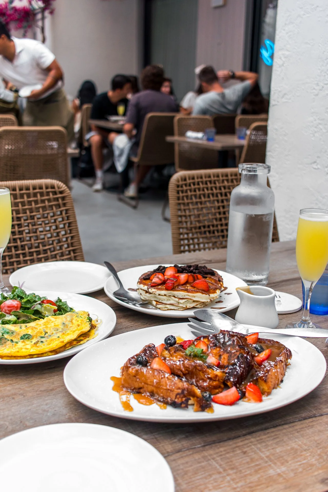 Plate of food with pancakes topped with strawberries, a small pitcher of syrup, a plate with an omelet and salad, a glass of water, a glass of orange juice, and a jug of water on the table in a restaurant with people dining in the background.