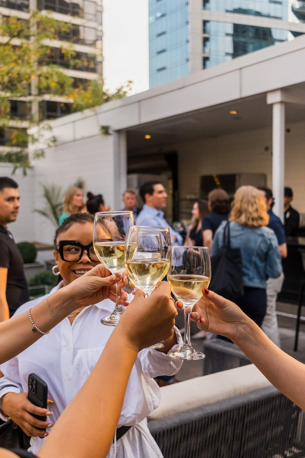 People raising glasses of white wine at a social gathering outdoors, with a woman smiling in the background and other guests in the scene.