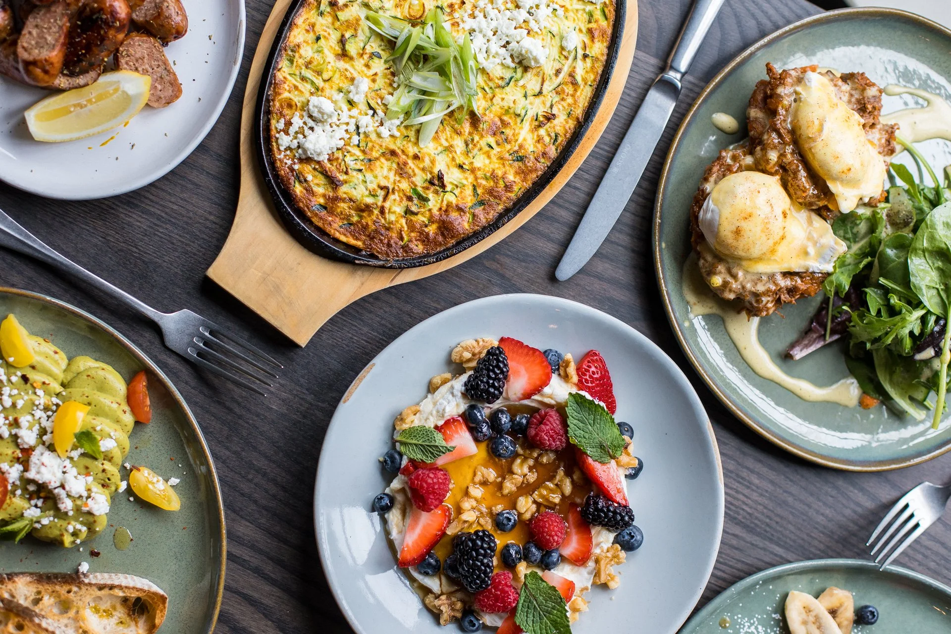 A variety of dishes on a dark wooden table, including a vegetable frittata with green onions and feta cheese in the center, eggs Benedict with hollandaise sauce and salad, a plate with fresh berries, strawberries, blueberries, raspberries, and a dessert with whipped cream, strawberries, blueberries, blackberries, walnuts, and honey.