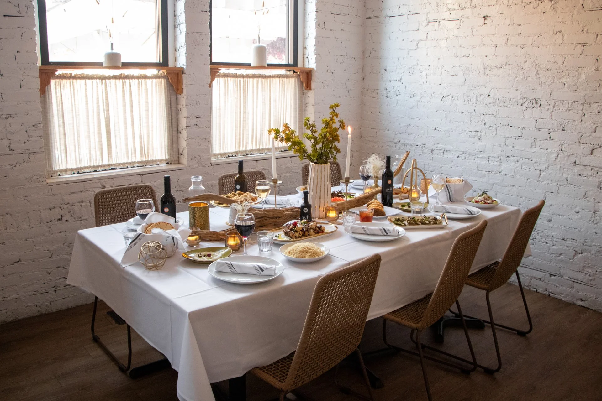 A dining table set for a meal with white tablecloth, plates, wine glasses, wine bottles, candles, and a large vase with flowers in a bright room with white brick walls and windows.