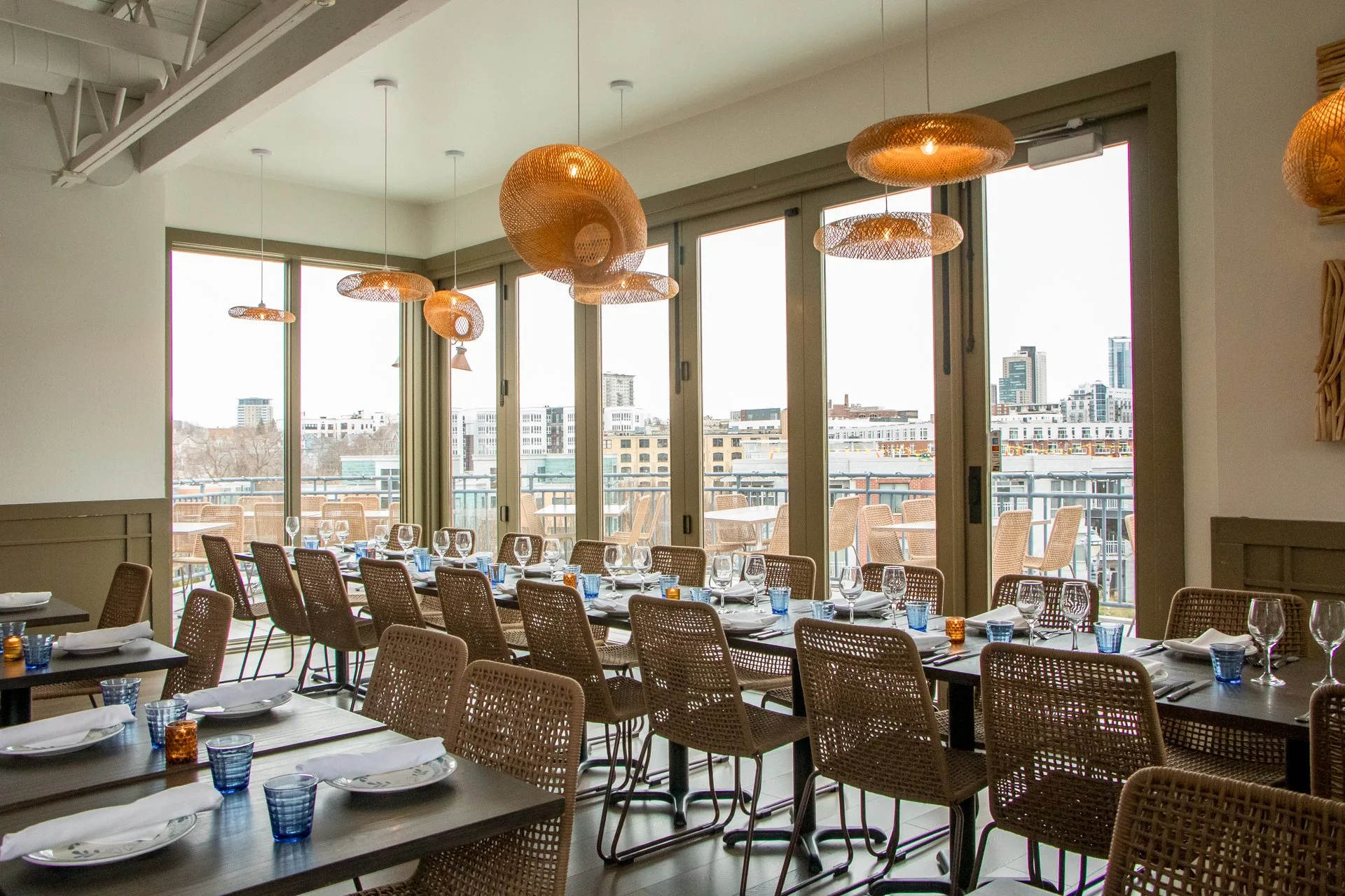 Restaurant dining area with tables set with white plates, silverware, wine glasses, and blue glasses, near large windows with cityscape view, featuring woven pendant lights hanging from the ceiling.
