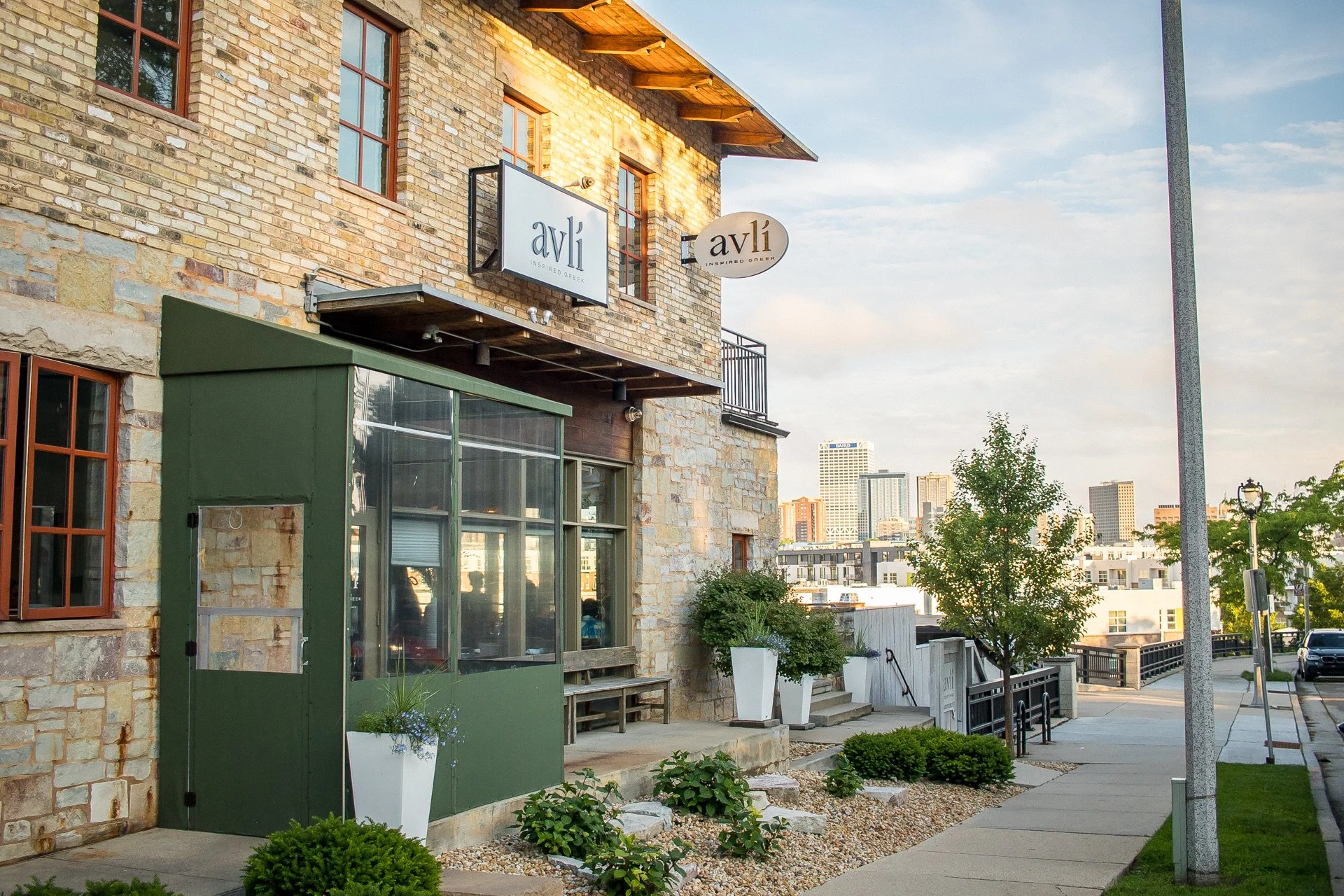 Exterior view of a restaurant or cafe named 'avli' with signs, a brick building, and outdoor decor including potted plants, a sidewalk, and city skyline in the background.