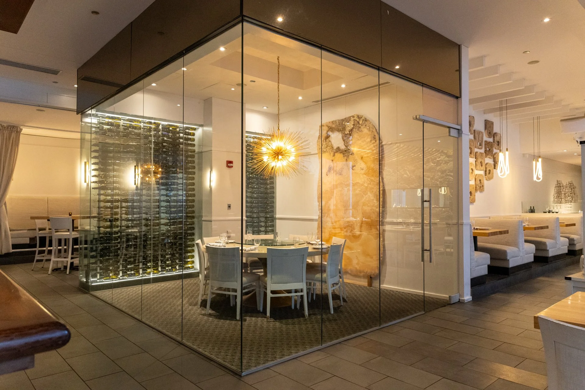 Interior of a modern restaurant section with a glass-walled private dining area, round dining table set with glasses and tableware, white chairs, wine cellar with wine bottles, wall art, and contemporary lighting fixtures.