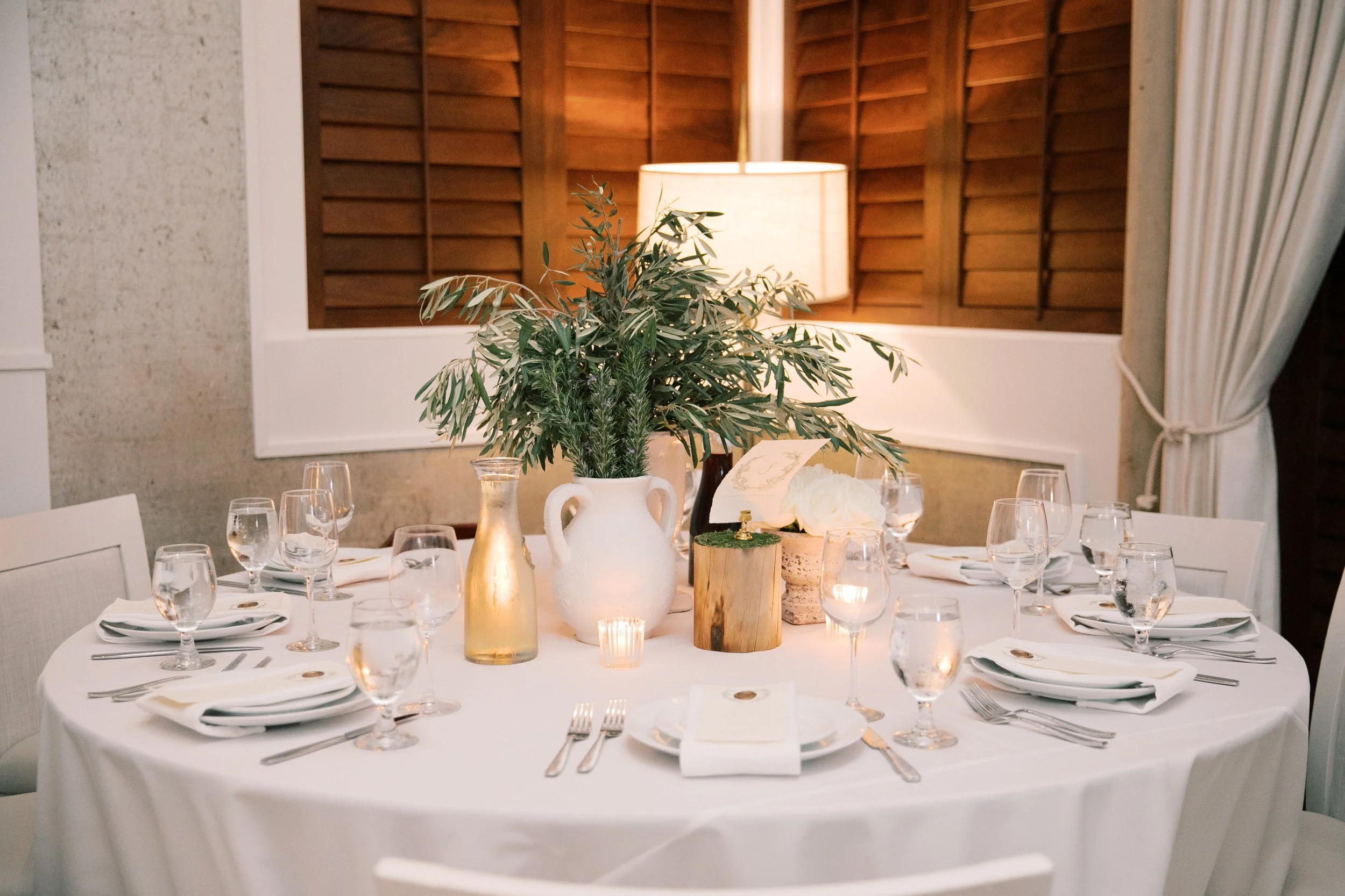 Round banquet table set for dinner with white tablecloth, multiple wine glasses, and plates with napkins. Centerpiece includes a large white vase with green leafy foliage, a smaller gold-colored vase, a wood log with a candle, and a small candle hold