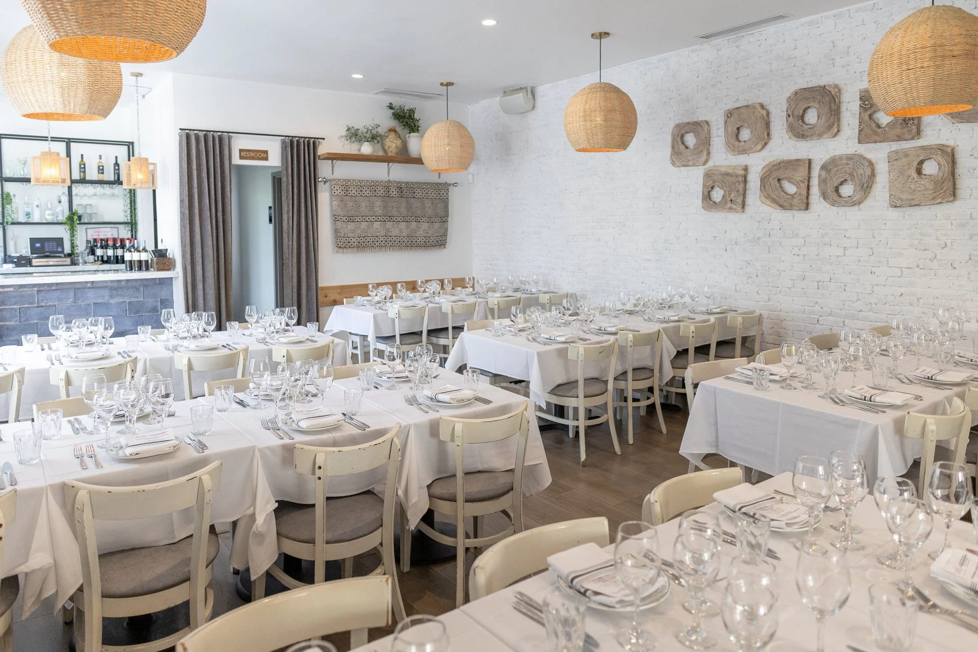 Interior of a restaurant with multiple white tables and chairs, set with wine glasses, water glasses, and napkins. Warm pendant lights hanging from the ceiling, large windows with curtains, and a small bar area in the background.