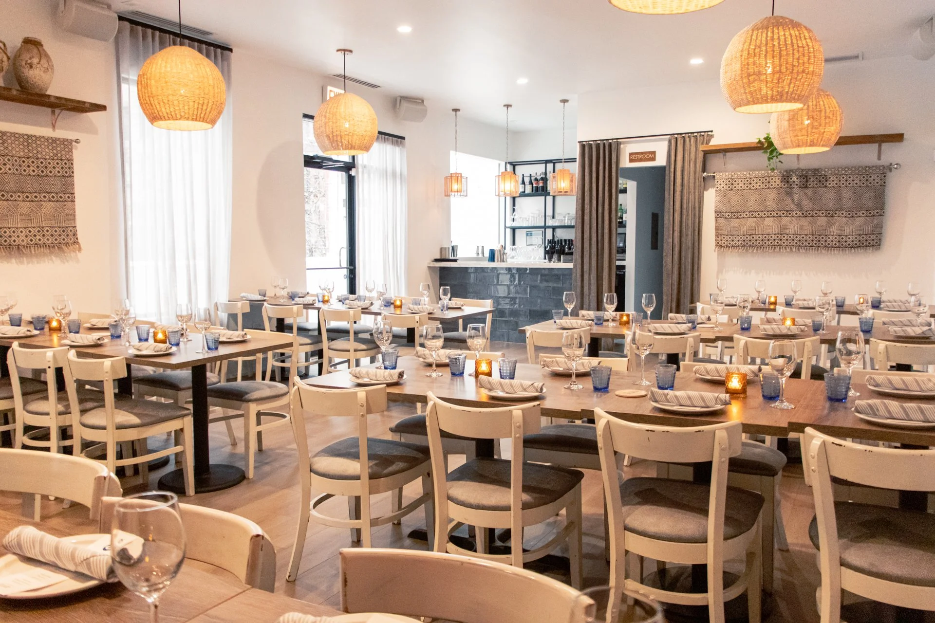 A cozy restaurant interior with multiple tables set with wine glasses, blue water glasses, and folded napkins, decorated with small candles, and with warm hanging lights and natural light coming through windows.