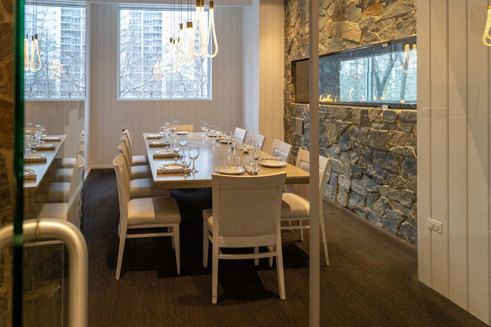 Empty dining room with a long wooden table set with plates, napkins, wine glasses, and utensils, and white chairs around it. Large windows with a snowy cityscape outside, stone accent wall with a narrow fireplace, and contemporary chandeliers hanging from the ceiling.