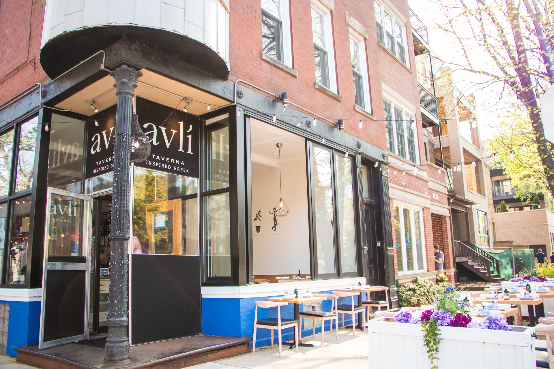 Exterior view of Avli, a Greek-inspired tavern and restaurant, with outdoor seating and string lights, on a sunny day with trees and residential buildings nearby.