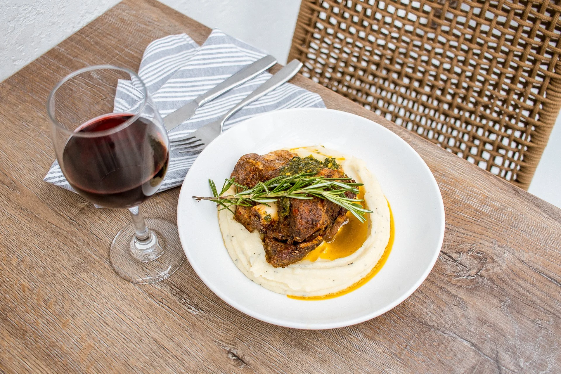 A plate of cooked beef topped with herbs, served with mashed potatoes and gravy, accompanied by a glass of red wine on a wooden table.