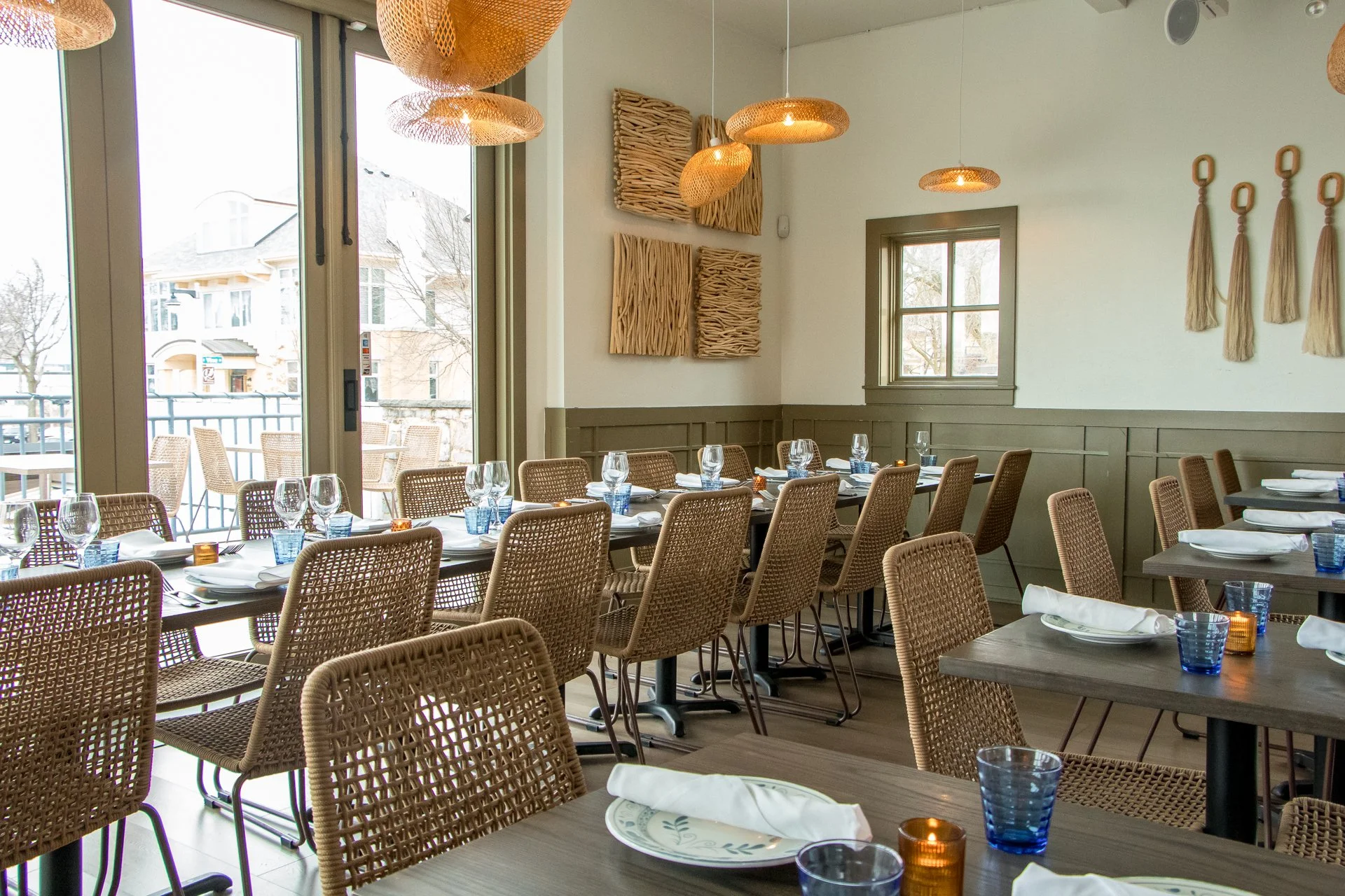 Interior of a restaurant with tables set and wicker chairs, large windows, and decor including woven wall art and hanging tassels.