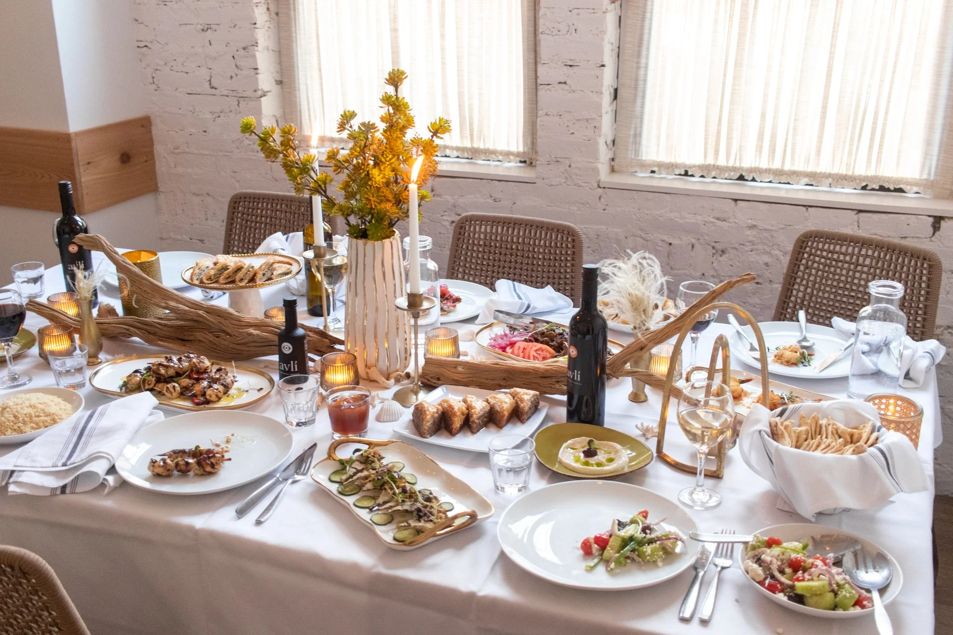 A dining table set for a meal with various plates of food, drinks, candles, and a centerpiece with flowers in a white striped vase in a bright room with large windows and white brick walls.