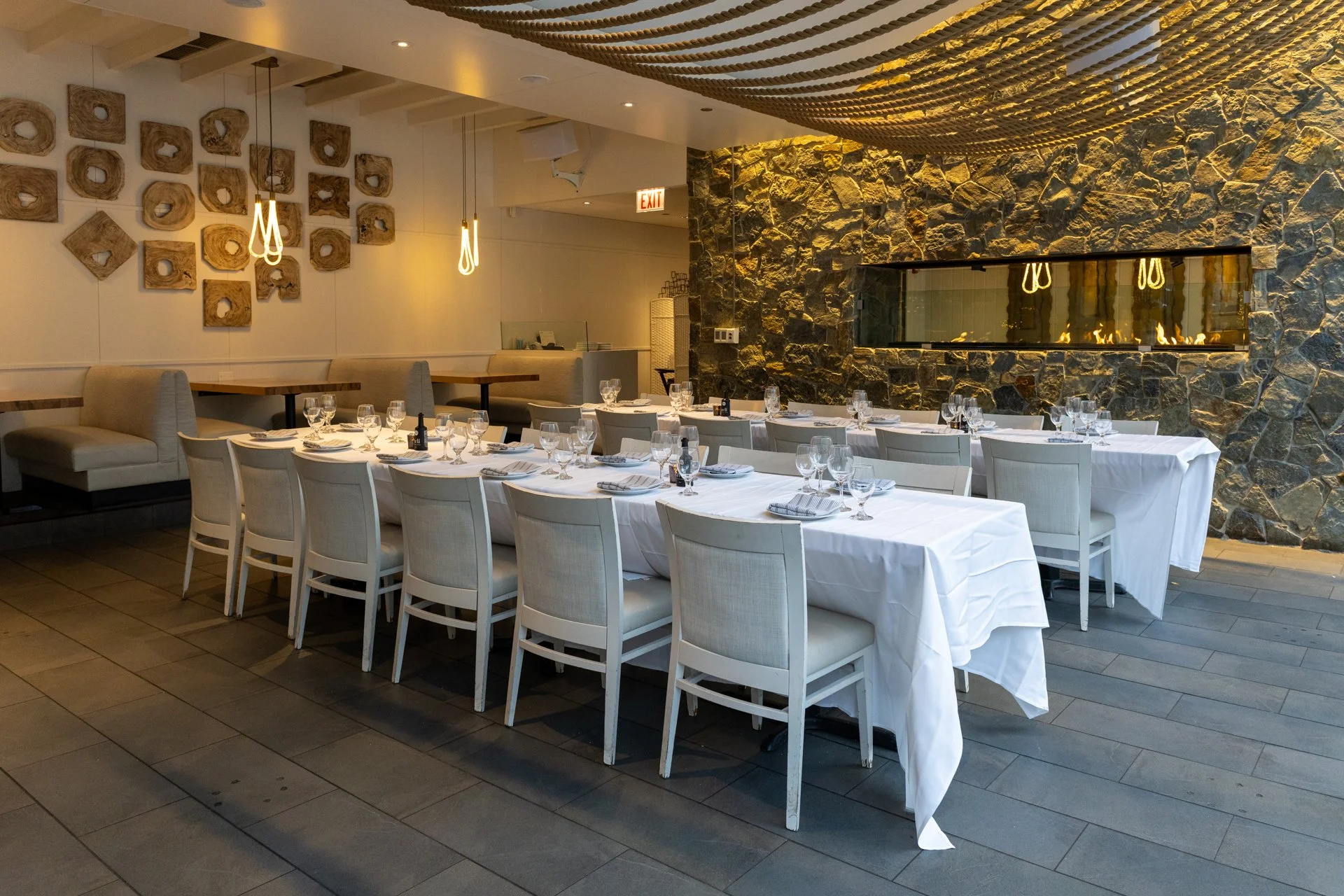 Elegant restaurant dining area with a long white table set with glasses and napkins, surrounded by white chairs, with a stone accent wall, modern pendant lights, and decorative wall art.