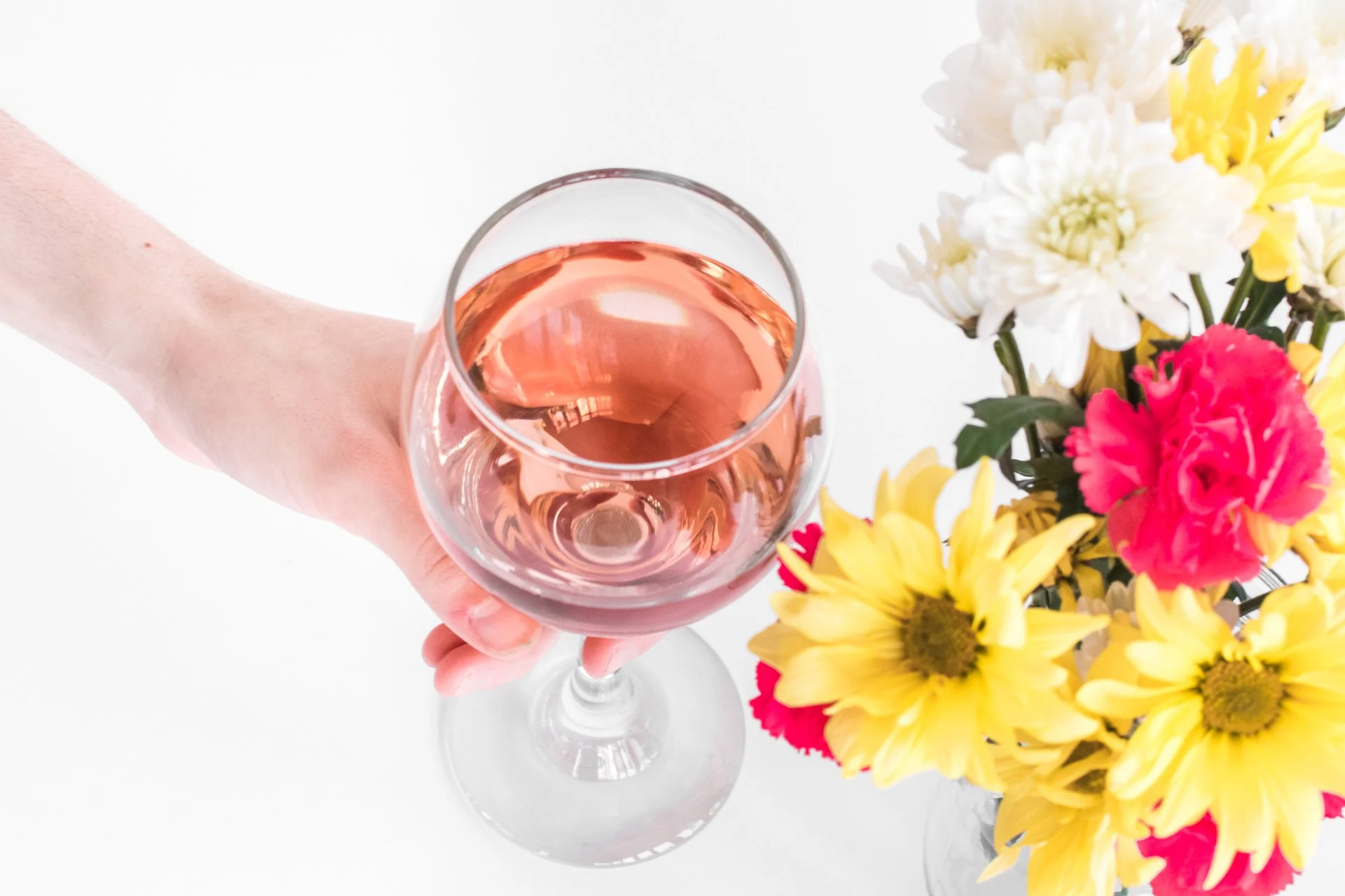 A hand holding a glass of rosé wine next to a colorful bouquet of white, yellow, and pink flowers on a white background.