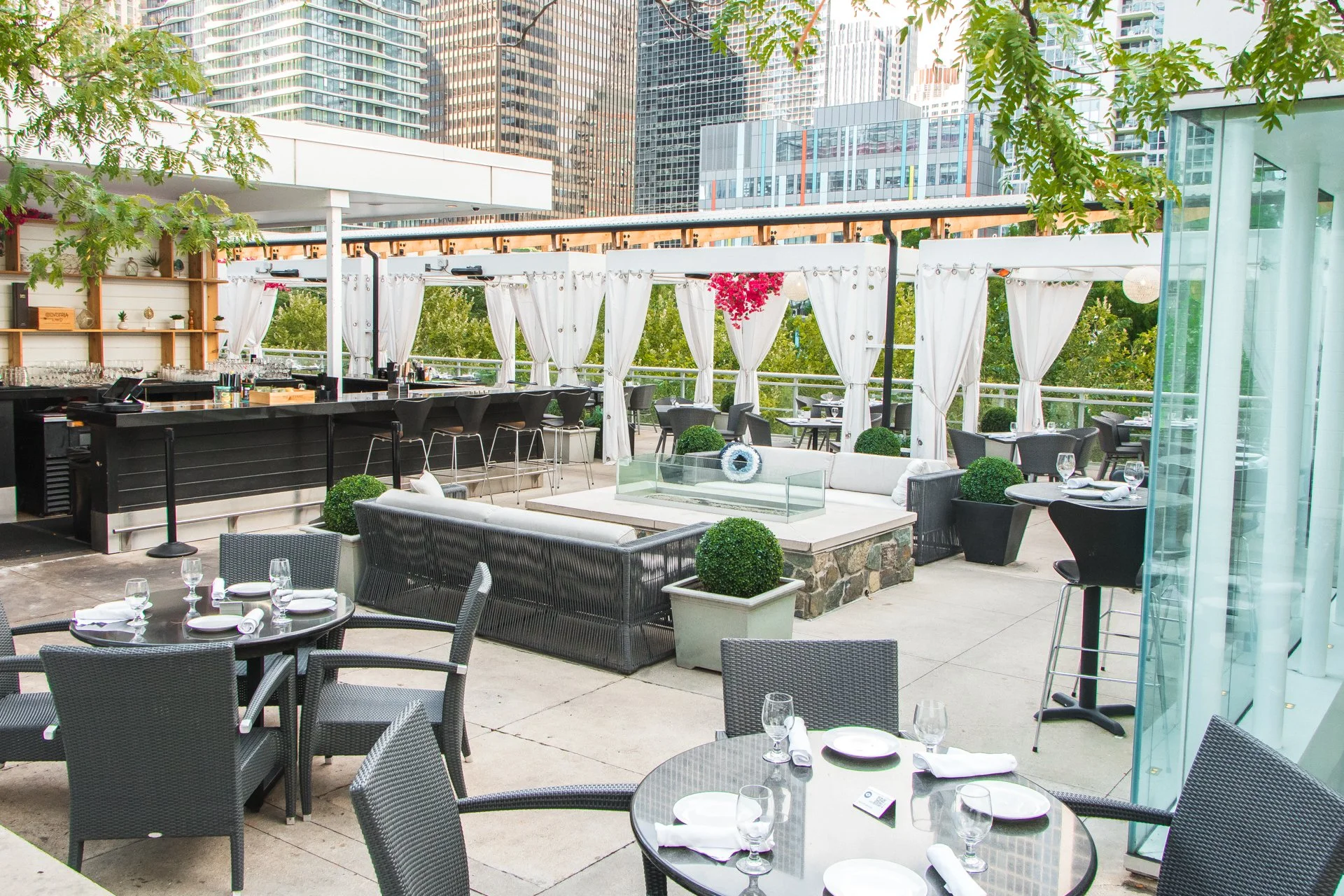 Outdoor rooftop restaurant with black tables and chairs, set with plates, glasses, and napkins. There is a bar area with high stools, white curtains, potted plants, trees, and city skyscrapers in the background.