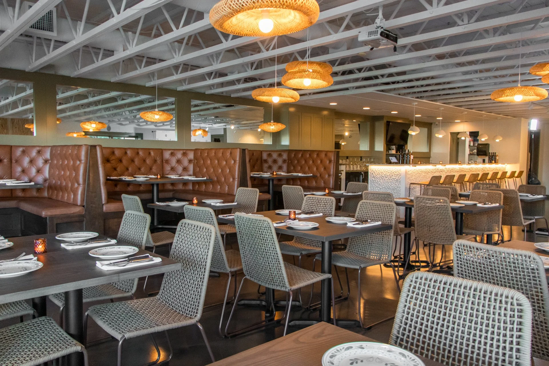 Empty restaurant dining area with brown leather booths, tables set with plates and utensils, wicker chairs, and warm lighting fixtures hanging from the ceiling.