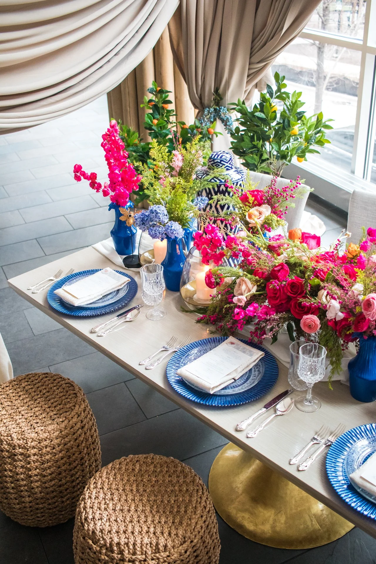 A table set for a meal with blue plates, crystal glasses, and silverware, decorated with colorful flower arrangements and candles, near a window with curtains.