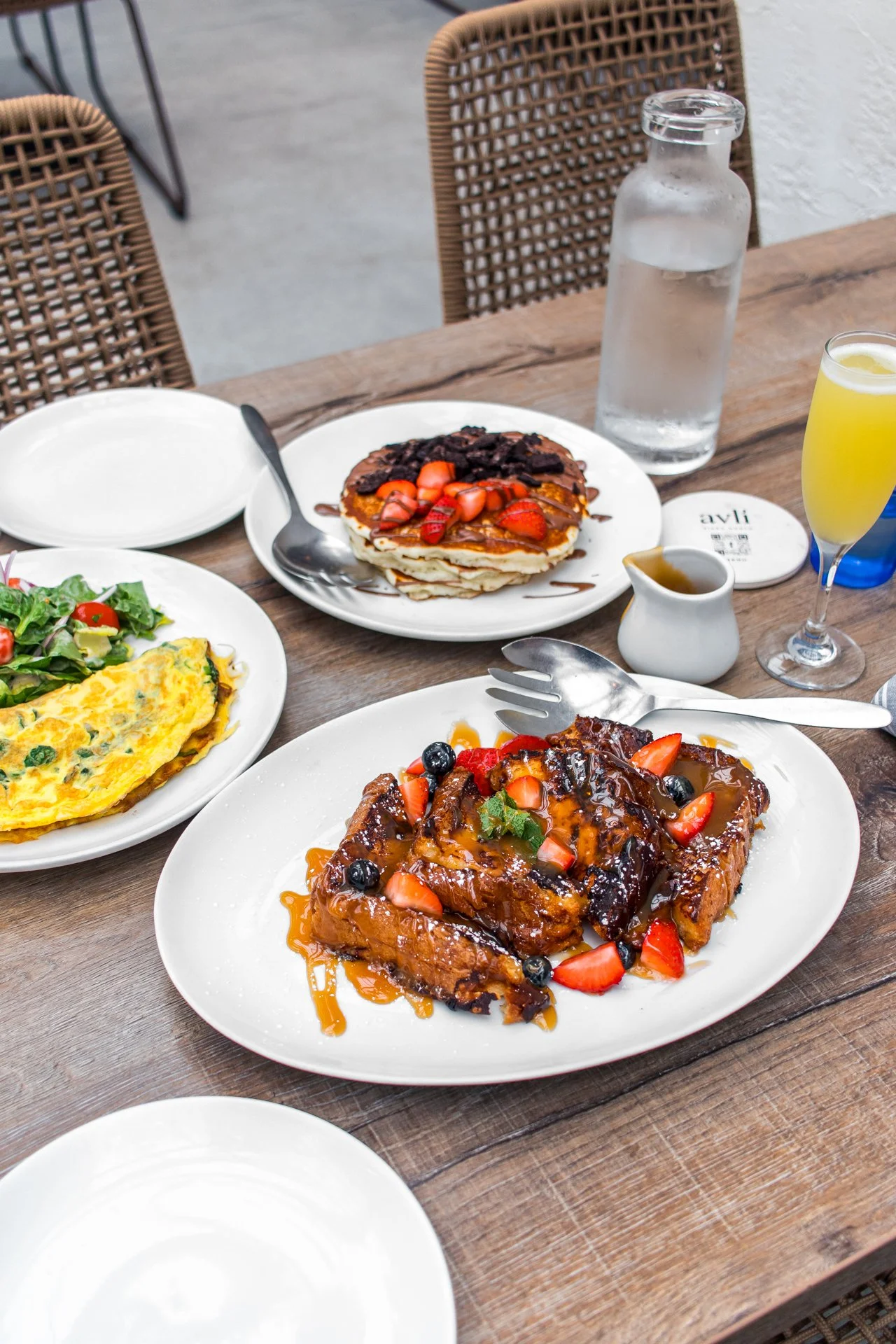 A breakfast table with various dishes including a plate of French toast topped with strawberries and chocolate, another plate of French toast with syrup and berries, an omelette with vegetables, a salad, a bottle of water, a glass of lemonade, and a small pitcher of syrup.