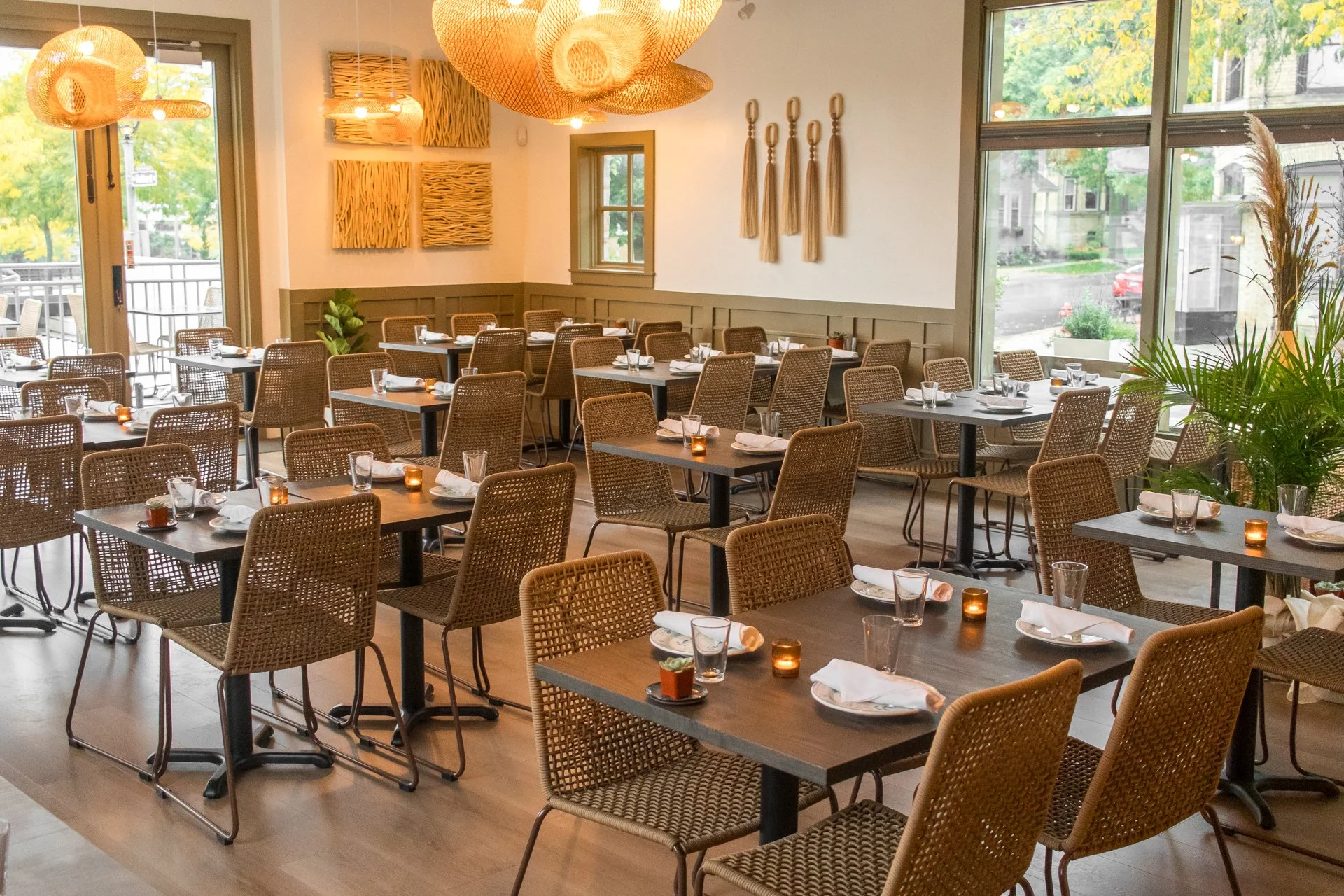 A restaurant with multiple tables set with white napkins, glassware, and small candles, featuring wicker chairs, indoor plants, and wall decorations, with large windows letting in natural light.