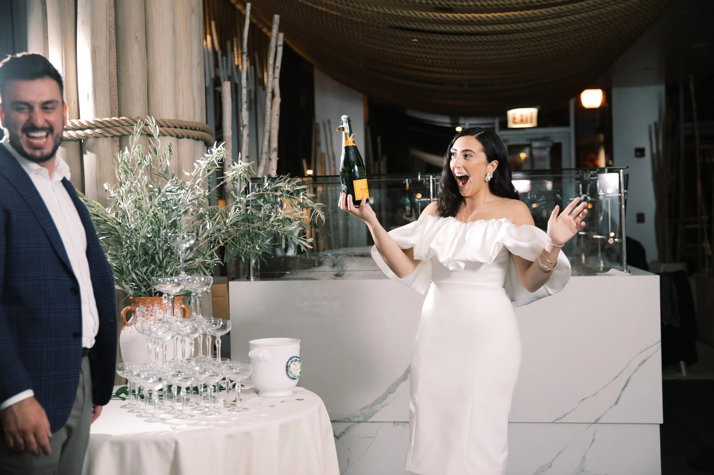 Woman in white dress celebrating with champagne at a social event inside a modern venue, with a man in a suit smiling nearby and a table with stacked champagne glasses and greenery decor.
