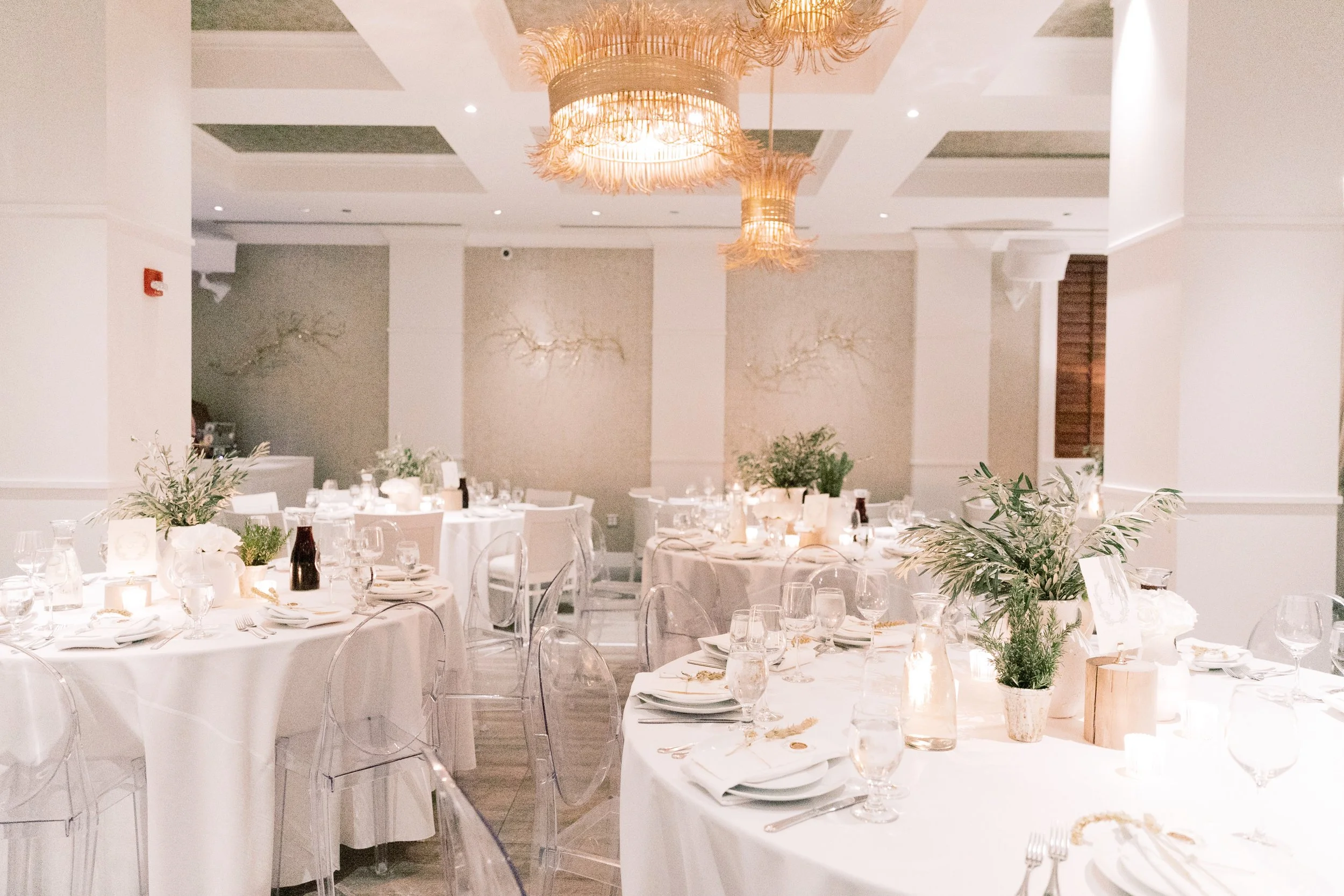 Elegant event hall with round tables decorated with white tablecloths, greenery centerpieces, candles, and glassware. Transparent chairs surround the tables. Soft ambient lighting and decorative wall art add to the sophisticated atmosphere.