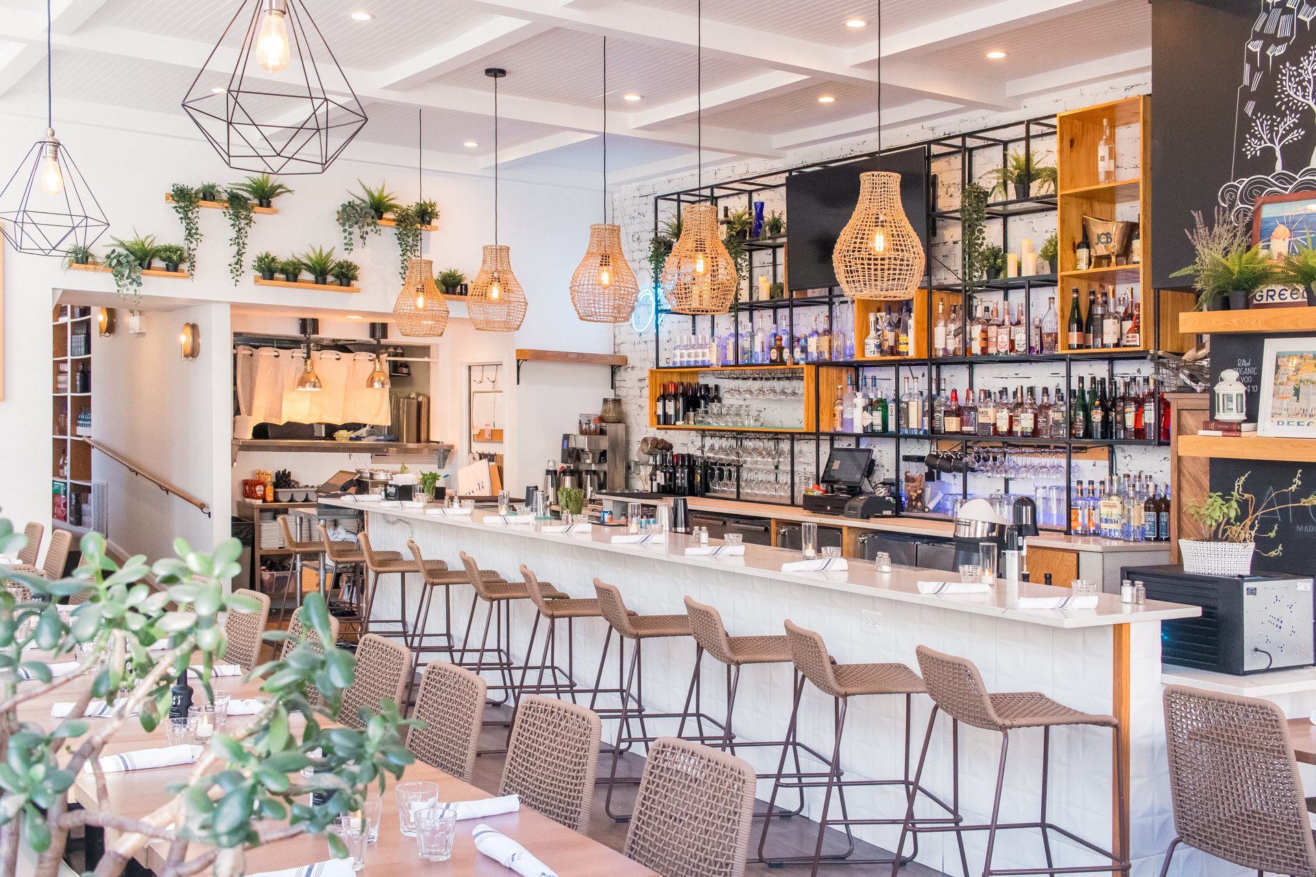 Interior of a modern bar and restaurant with a white marble counter, wicker bar stools, hanging pendant lights, shelves with bottles of alcohol, and plants for decor.