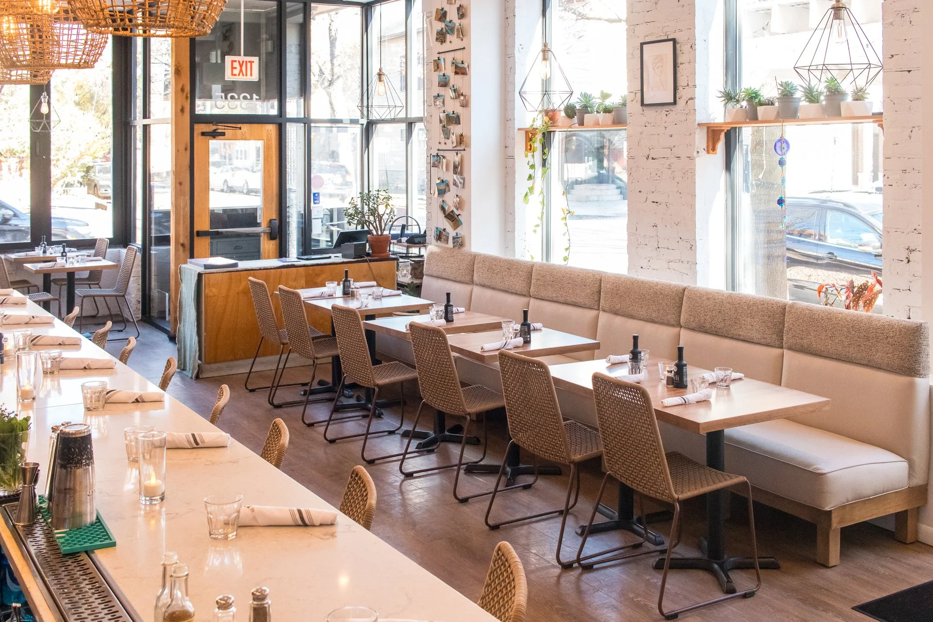 Empty restaurant with wooden and wicker furniture, white brick walls, large windows, plants, and modern light fixtures.