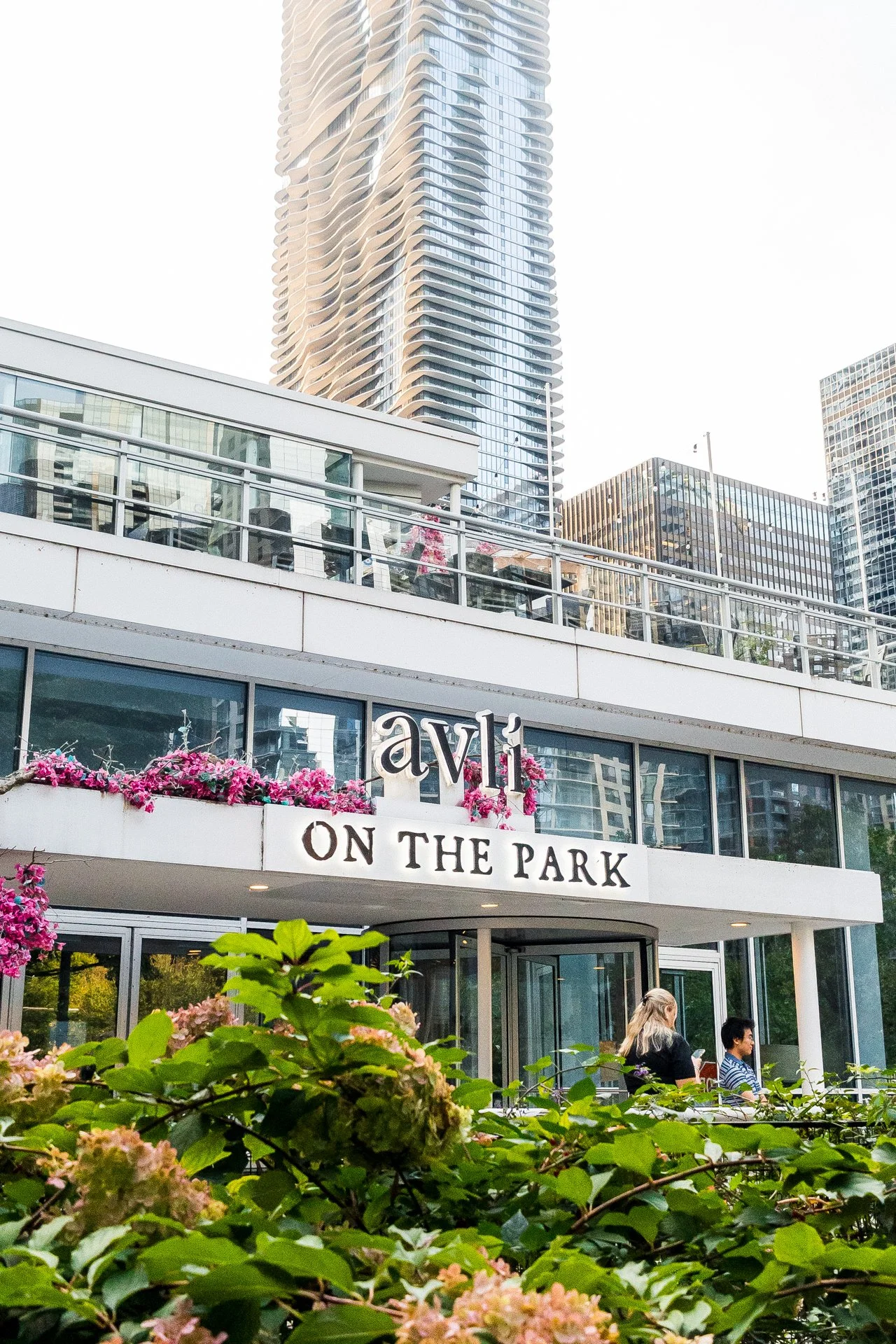 Outdoor view of Avli on the Park restaurant with pink flowers, greenery, modern glass buildings, and tall apartment or office skyscraper in the background in an urban setting