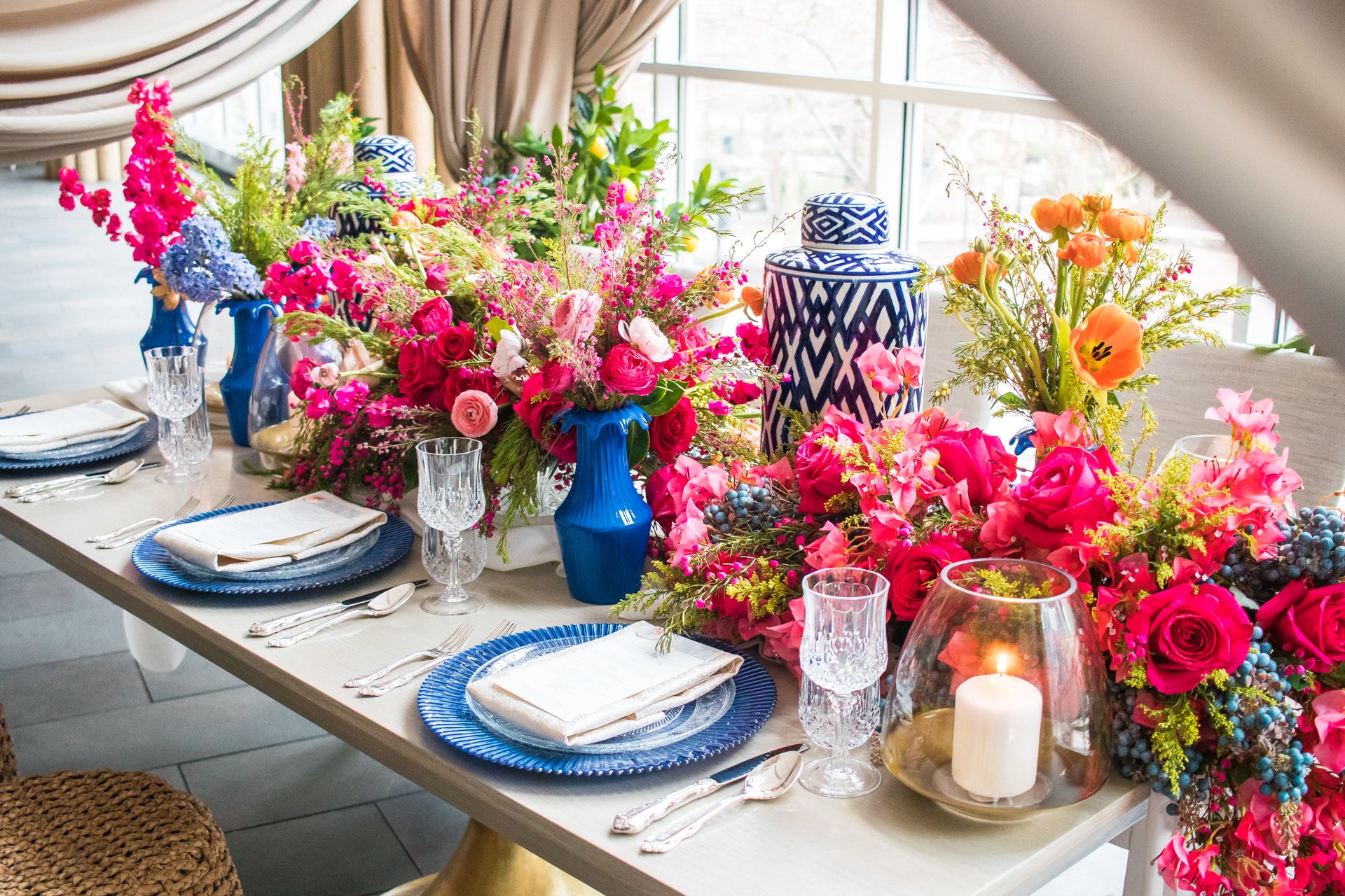 A table decorated with pink, red, and purple flowers in blue vases, set with blue plates, silverware, crystal glasses, and a candle inside a glass holder, with beige curtains and large windows in the background.