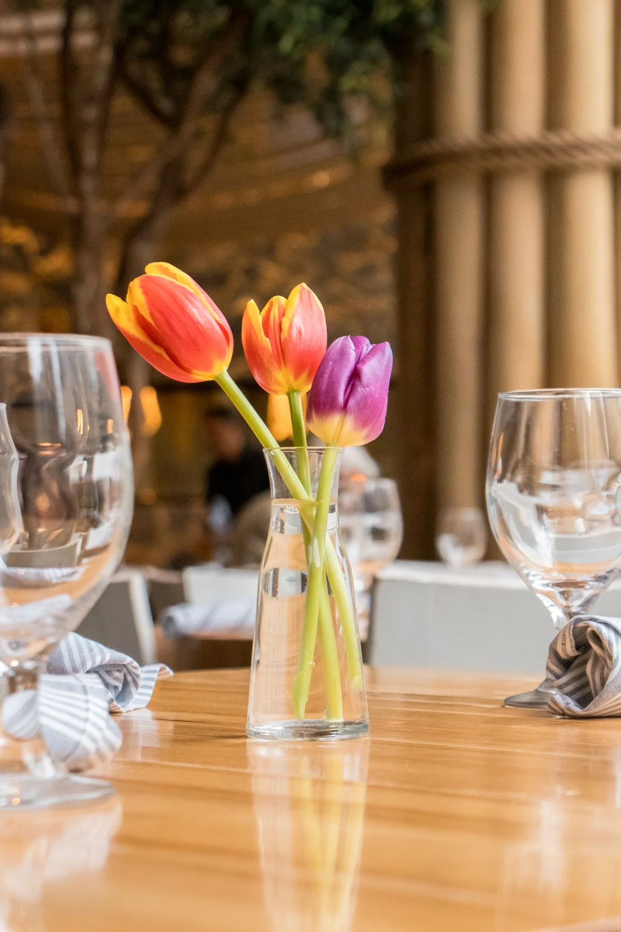 A small glass vase with three colorful tulips on a wooden table, surrounded by empty wine glasses in a restaurant or cafe setting.