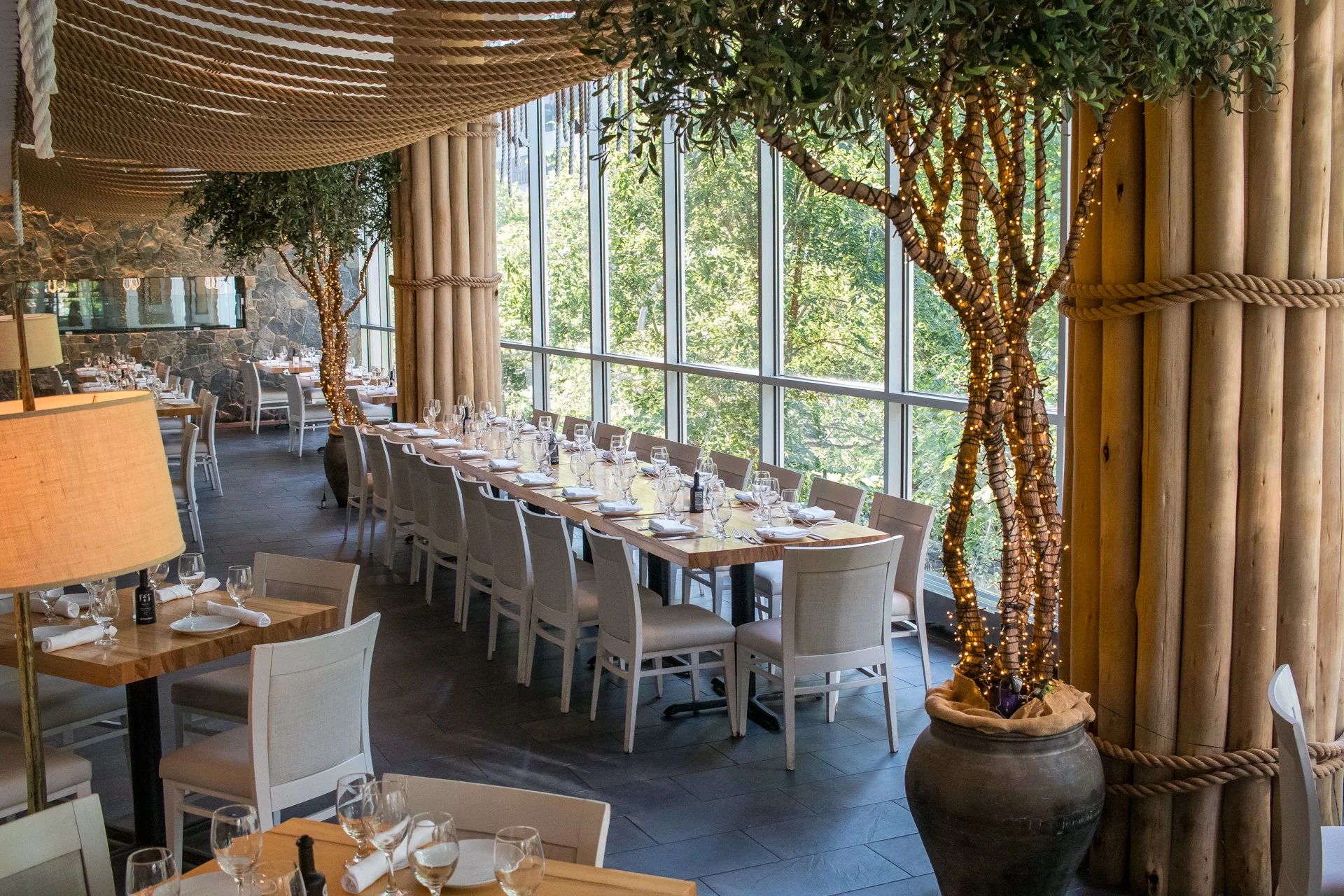 Elegant restaurant interior with long wooden tables set with wine glasses, plates, and napkins, large windows with greenery outside, decorative trees wrapped with string lights, and wood-paneled walls.