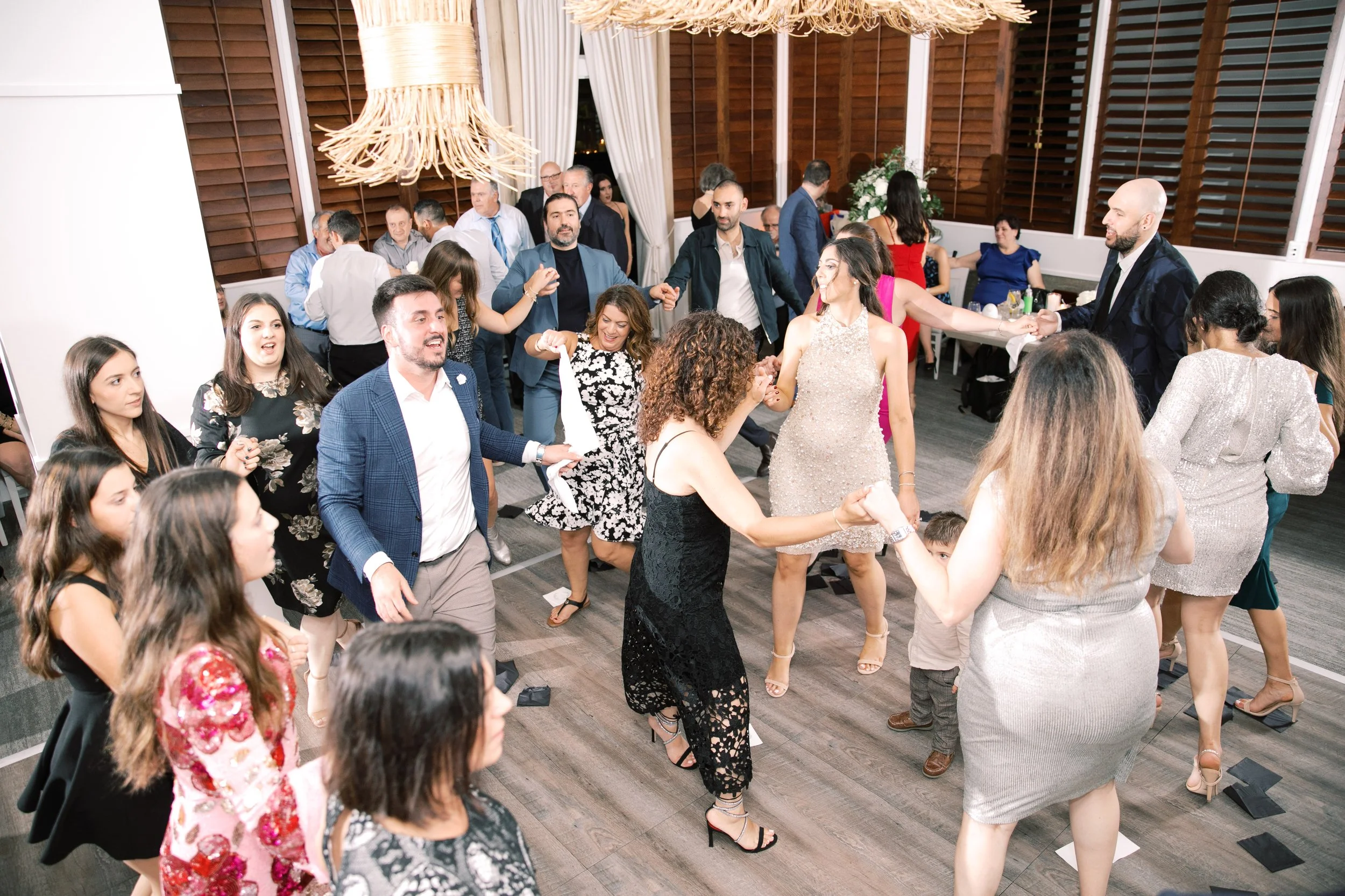 People dancing at a celebration or wedding reception in a decorated indoor venue with wooden walls and large windows with curtains.