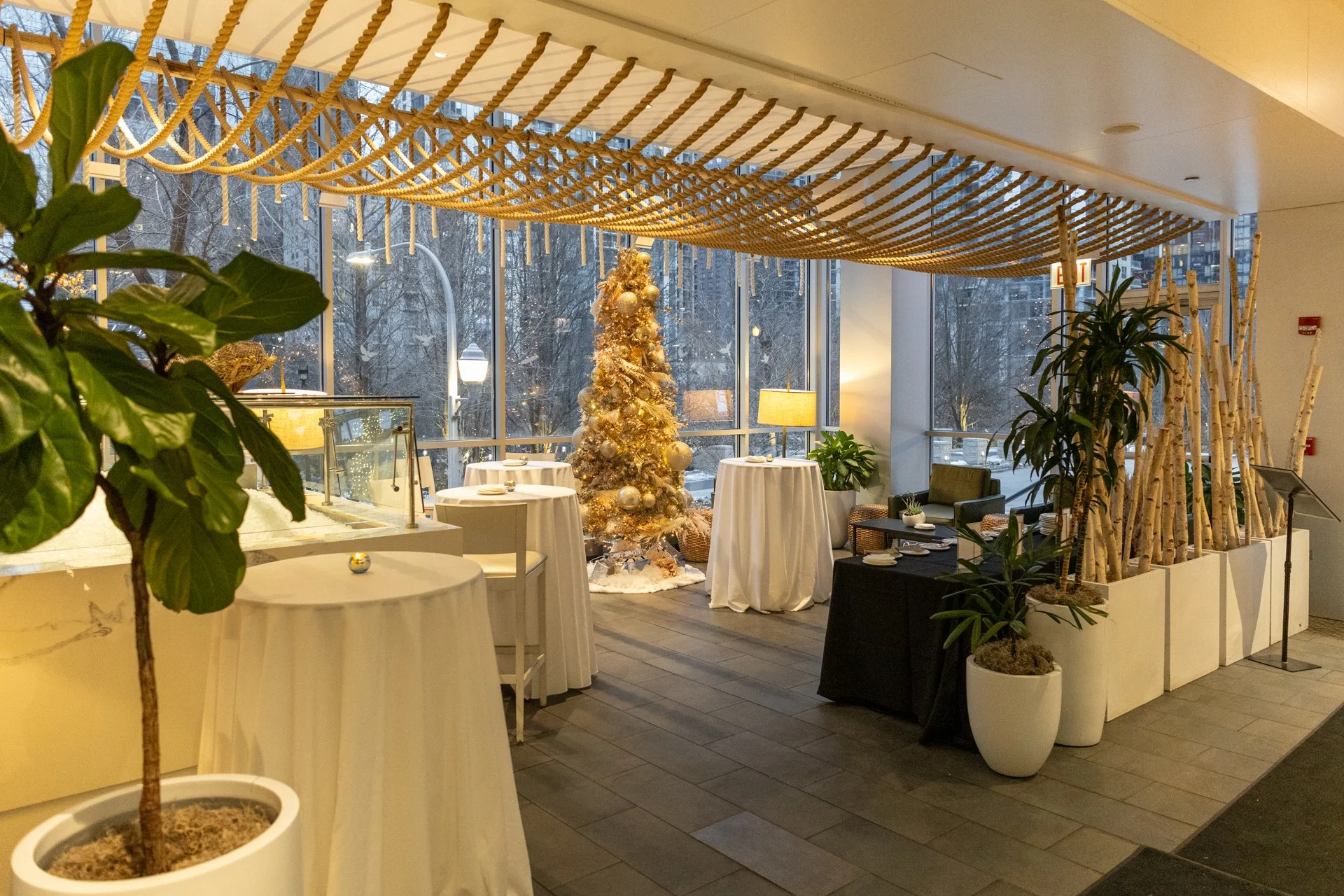 Indoor holiday decorated space with Christmas tree, tall potted plants, standing tables with white tablecloths, arrangement of birch branches in planters, large windows, and warm lighting.