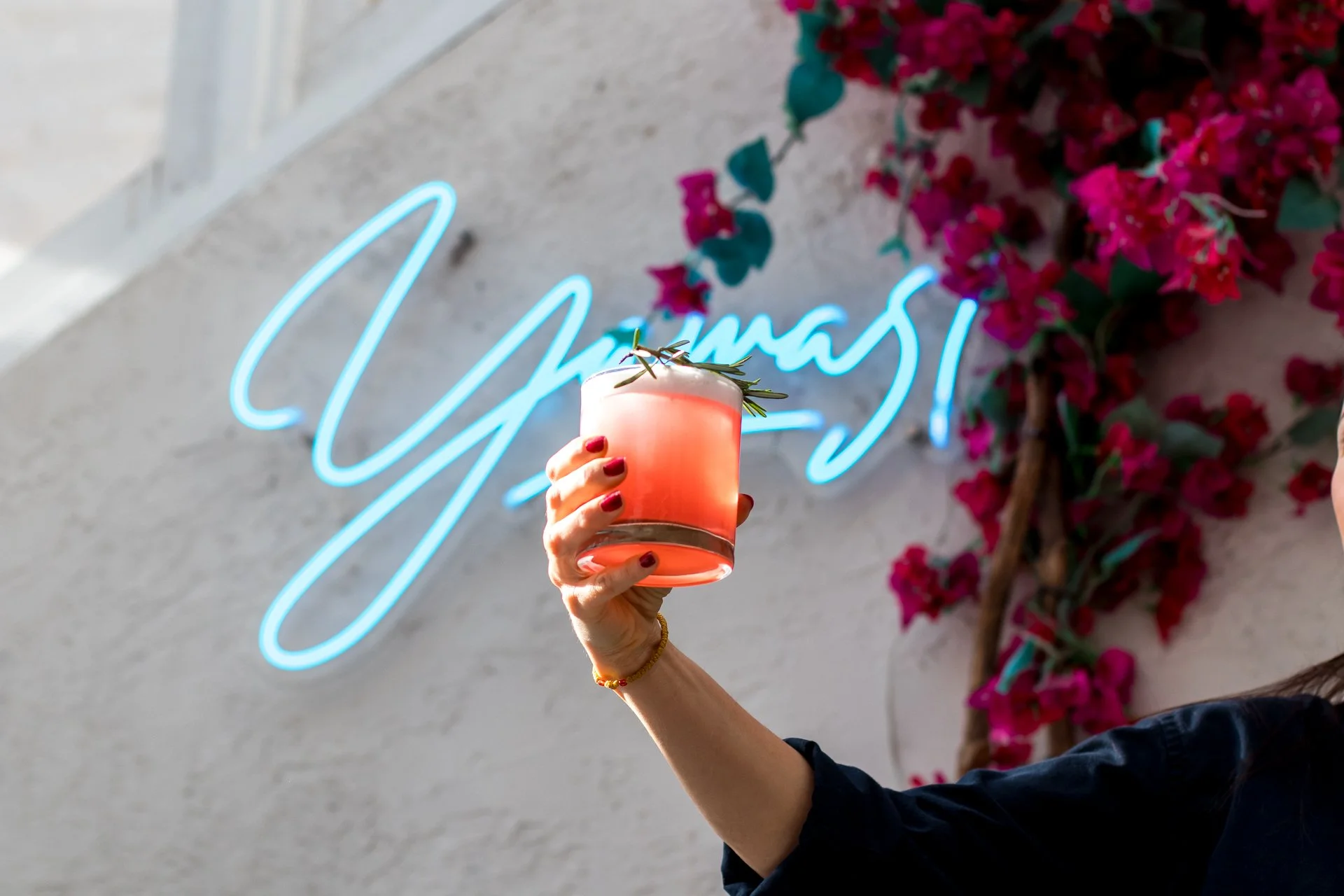 A person holding a pink drink garnished with a sprig of rosemary, with a neon sign reading 'Cheers!' in the background and pink flowers on the wall.
