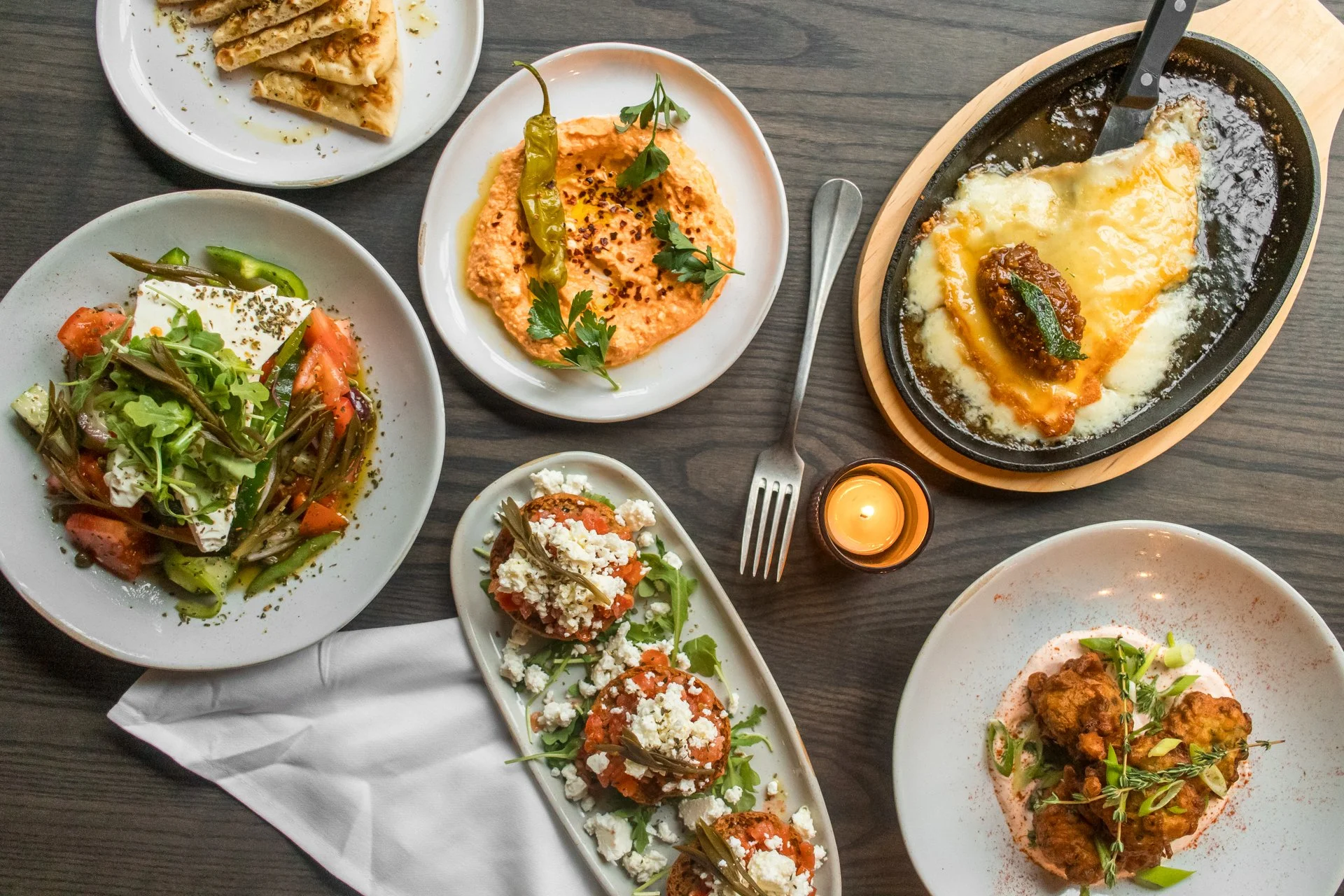 A variety of Middle Eastern dishes on a dark wooden table, including a plate of stuffed grape leaves, a salad with fresh vegetables and herbs, a dish of hummus topped with chili and parsley, a skillet of baked cheesy eggplant, and fried cauliflower with herbs and spices, with a fork, a small candle, and a white napkin.