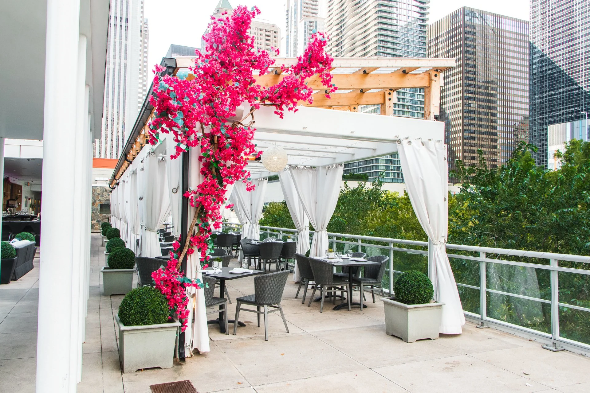 Empty outdoor rooftop restaurant with white curtains, black tables and chairs, pink flowering tree, potted plants, and city skyscrapers in the background.