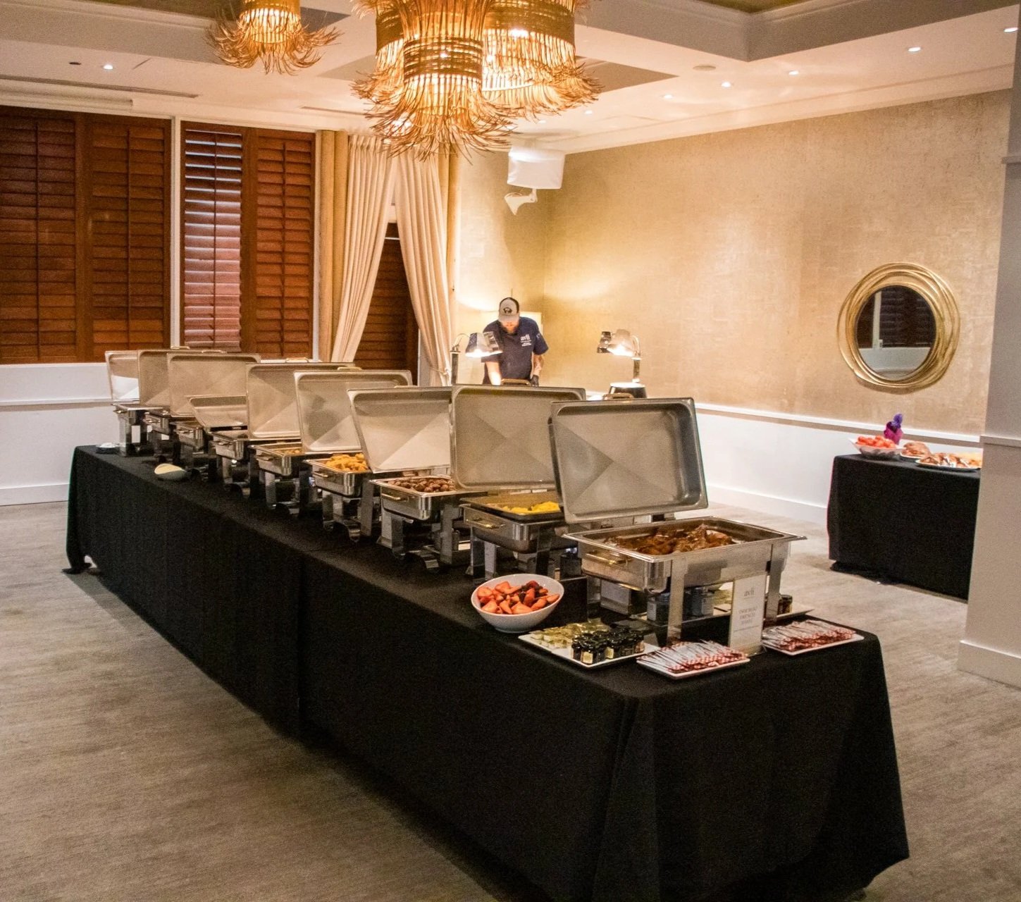 Buffet table with chafing dishes containing food, bowls of fruit, and condiments in a banquet hall with beige walls, curtains, wooden shutters, and decorative lighting.