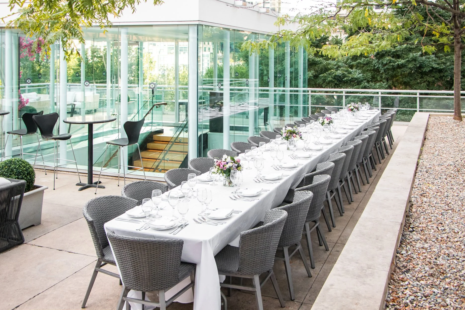 Long outdoor dining table set for a formal event with white tablecloths, glassware, white plates, silverware, and floral centerpieces, on a terrace with glass walls and greenery in the background.