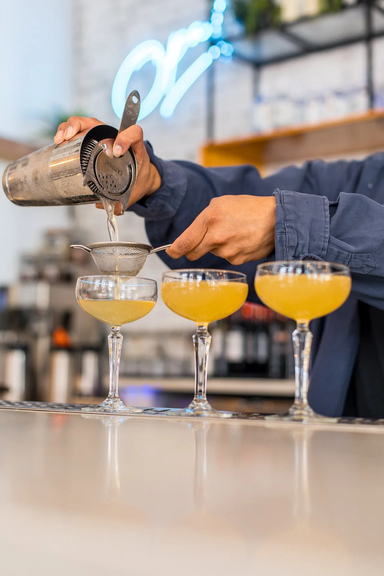 A bartender pouring a yellow cocktail through a strainer into coupe glasses.