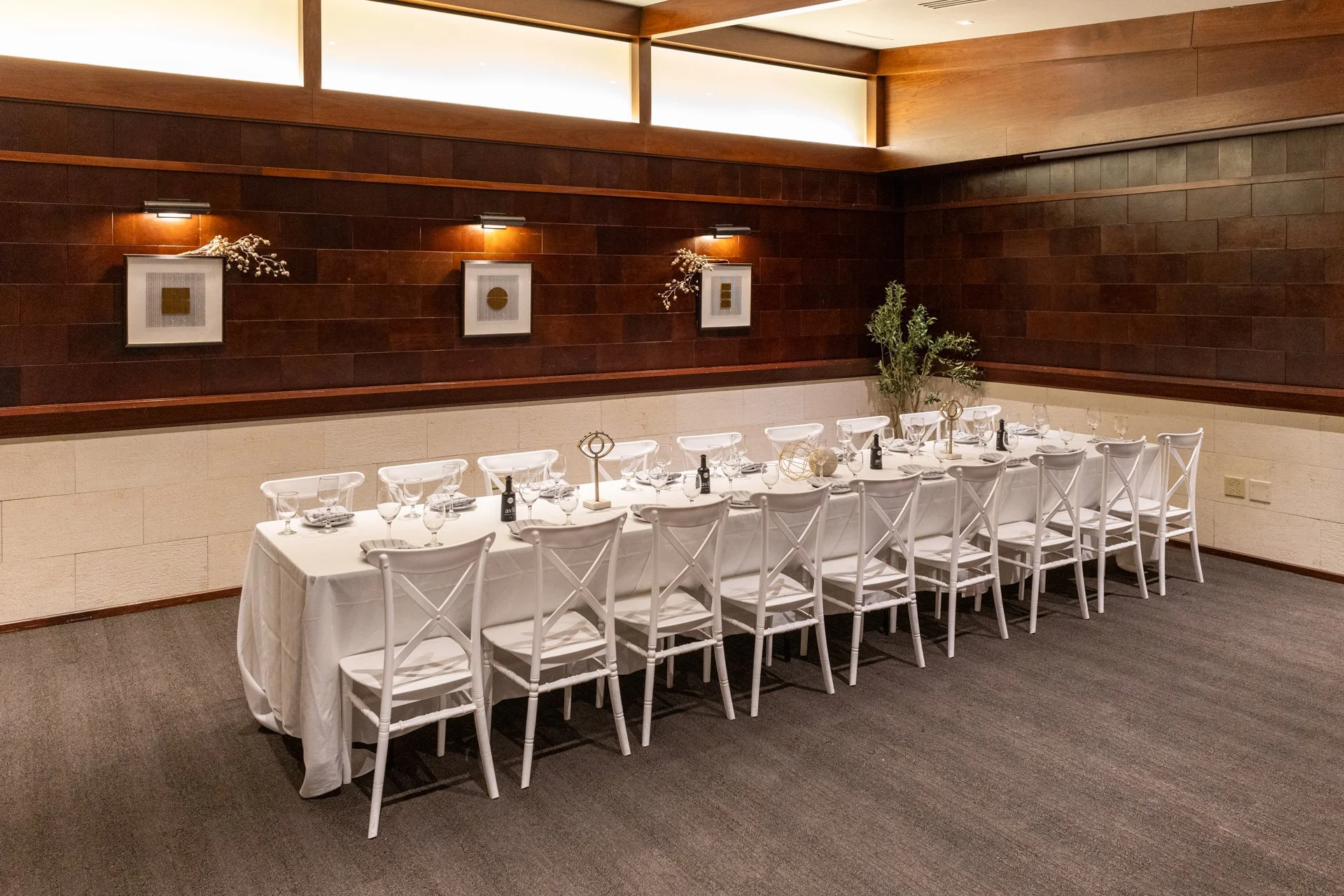 Long banquet table set for a formal event with white tablecloth, glasses, and small bottles, in a room with brown and beige walls, decorative artwork, and a potted plant.