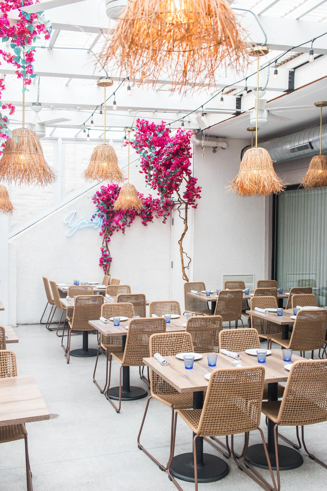 Indoor restaurant with wooden tables, woven chairs, and blue glasses set on each table. Pink flowering bougainvillea climbing the wall, hanging straw pendant lights, and string lights overhead create a cozy, tropical ambiance.