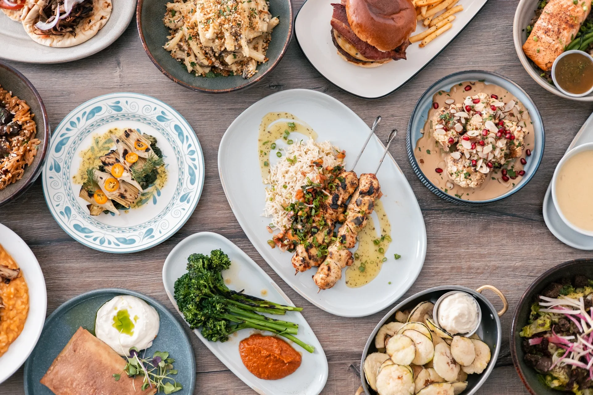 A variety of dishes on a wooden table, including grilled chicken skewers with rice, broccoli with sauce, pasta salad, salmon with greens, a burger, and other plated foods.