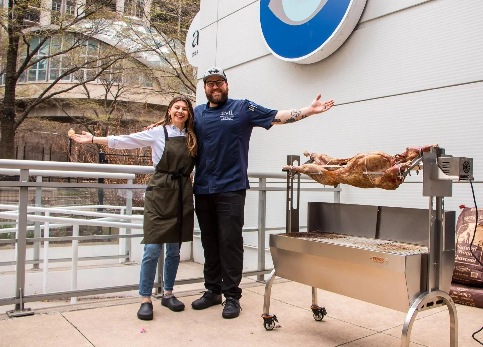 Two smiling people, a woman and a man, stand with arms open near a whole roasted animal on a spit, outside on a patio. The man wears a chef's uniform and the woman has a dark apron. There is a barbecue grill and a bag of charcoal in the background.