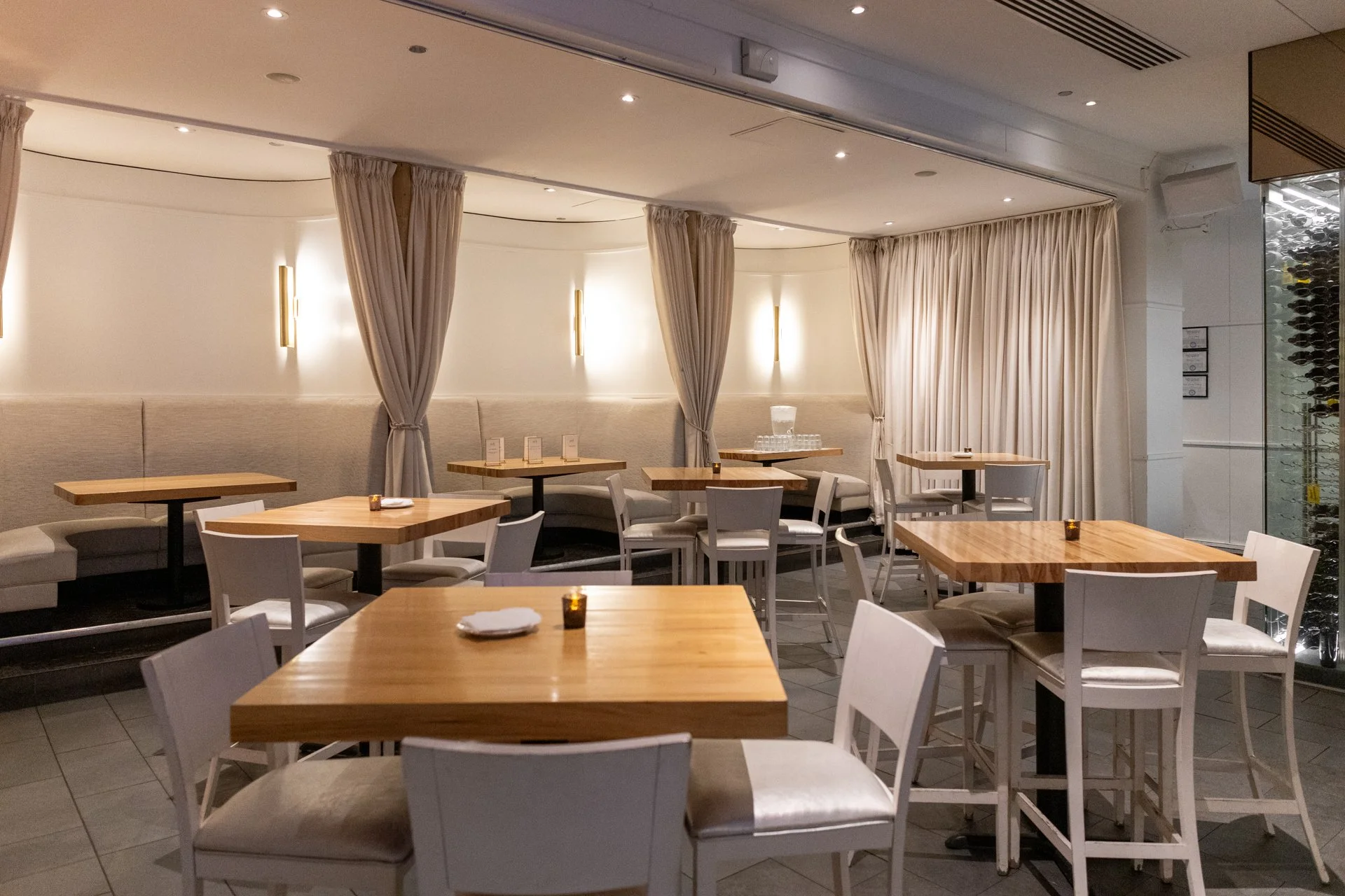 Empty modern restaurant with wooden tables, white chairs, beige curtains, and wall-mounted lights.