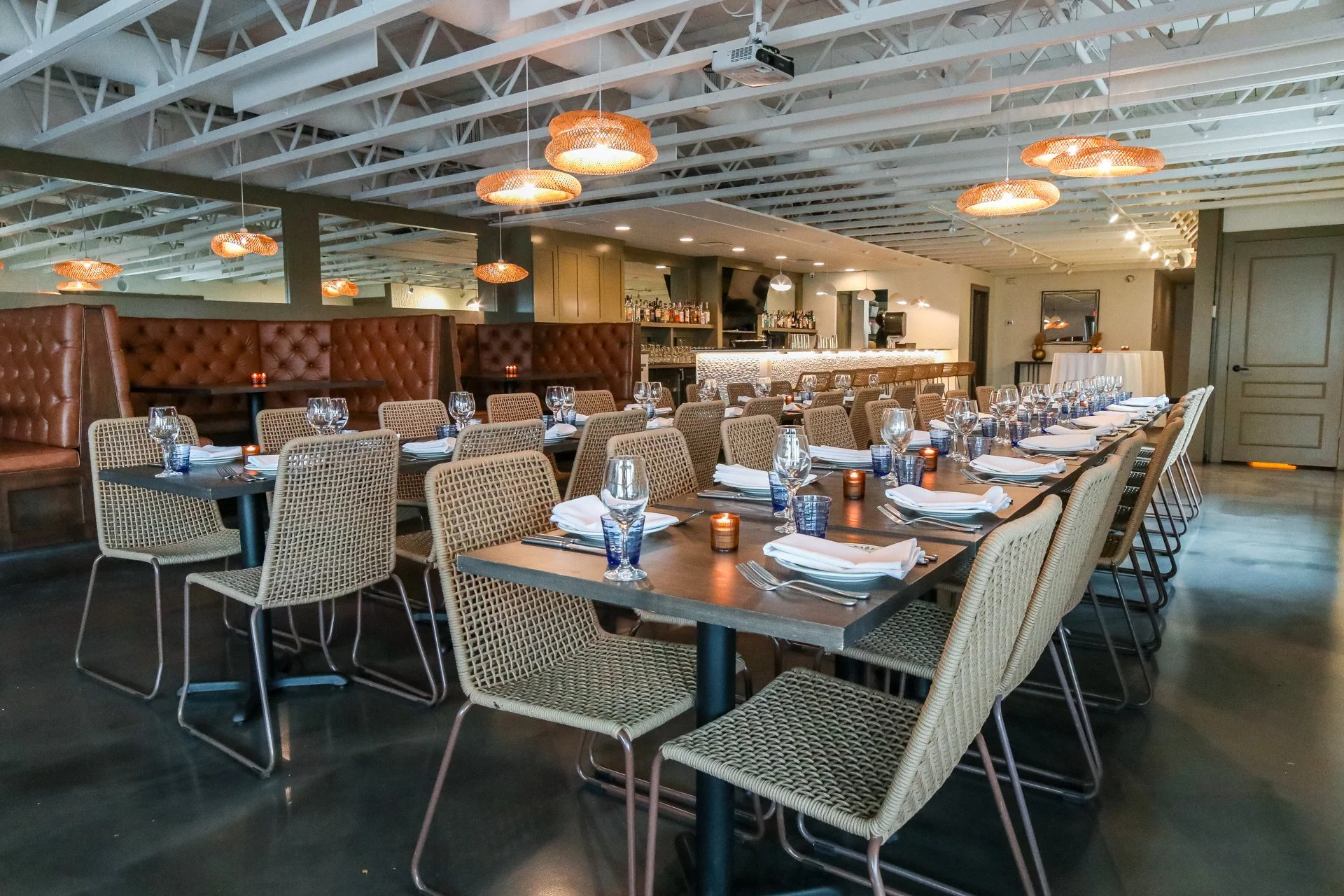 A modern restaurant interior with long dining tables set for a meal, with wine glasses, folded napkins, and candles, featuring a bar area in the background and warm ambient lighting.
