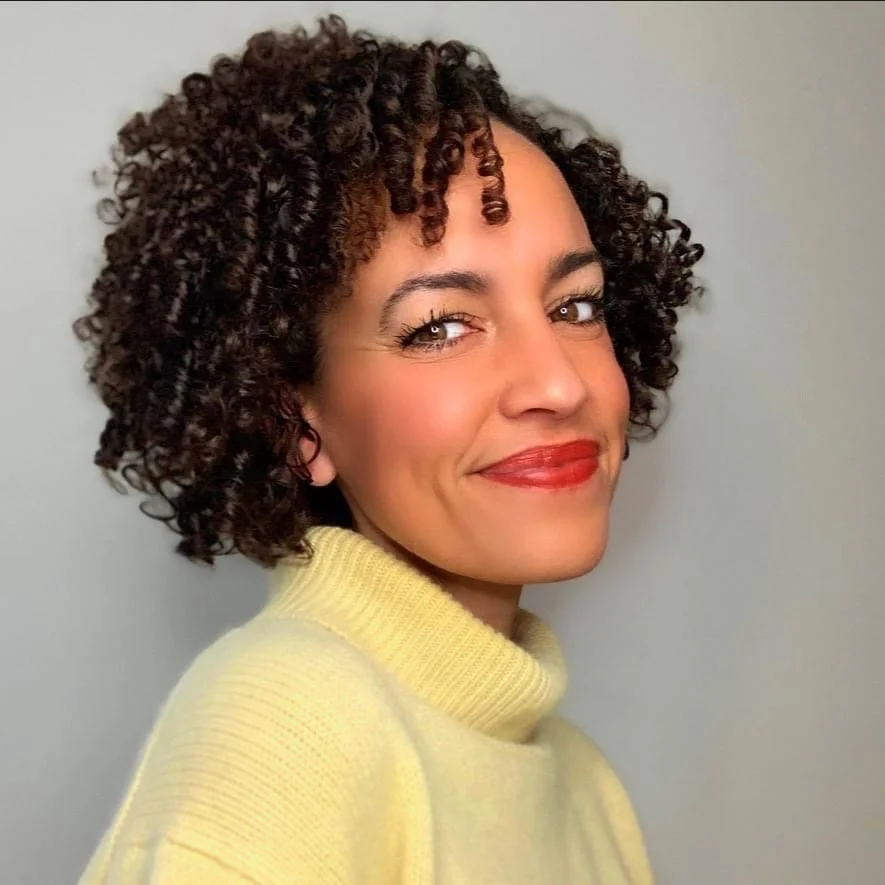 A woman with short, curly hair and red lipstick, smiling and looking at the camera, wearing a yellow turtleneck sweater against a plain background.