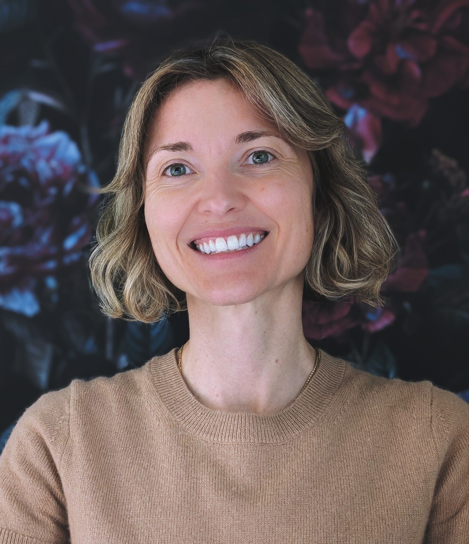 A woman with short light brown hair smiling, wearing a beige sweater, against a dark floral background.