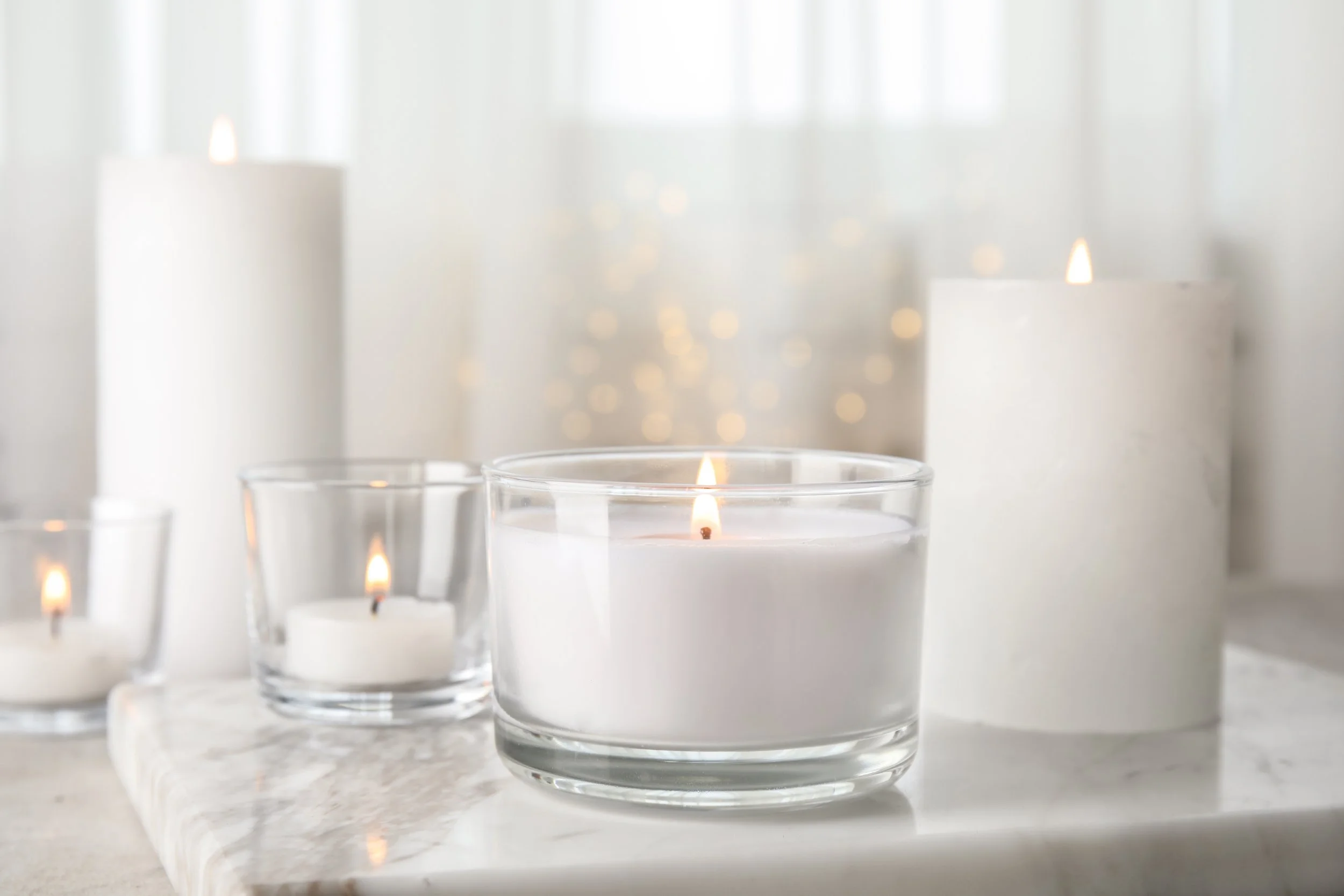 Different sizes of lit white candles arranged on a marble surface in a bright room.