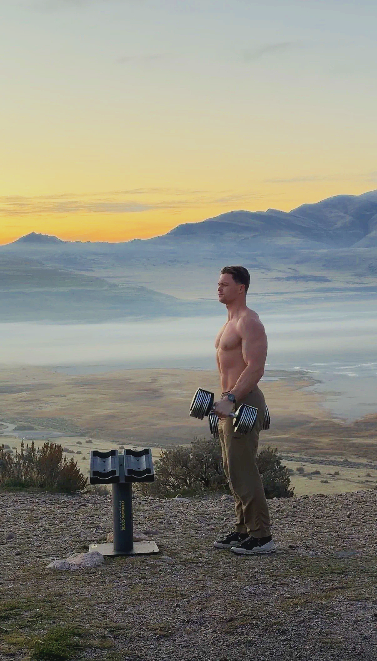 A shirtless man holding dumbbells stands outdoors during sunrise or sunset with mountains in the background.