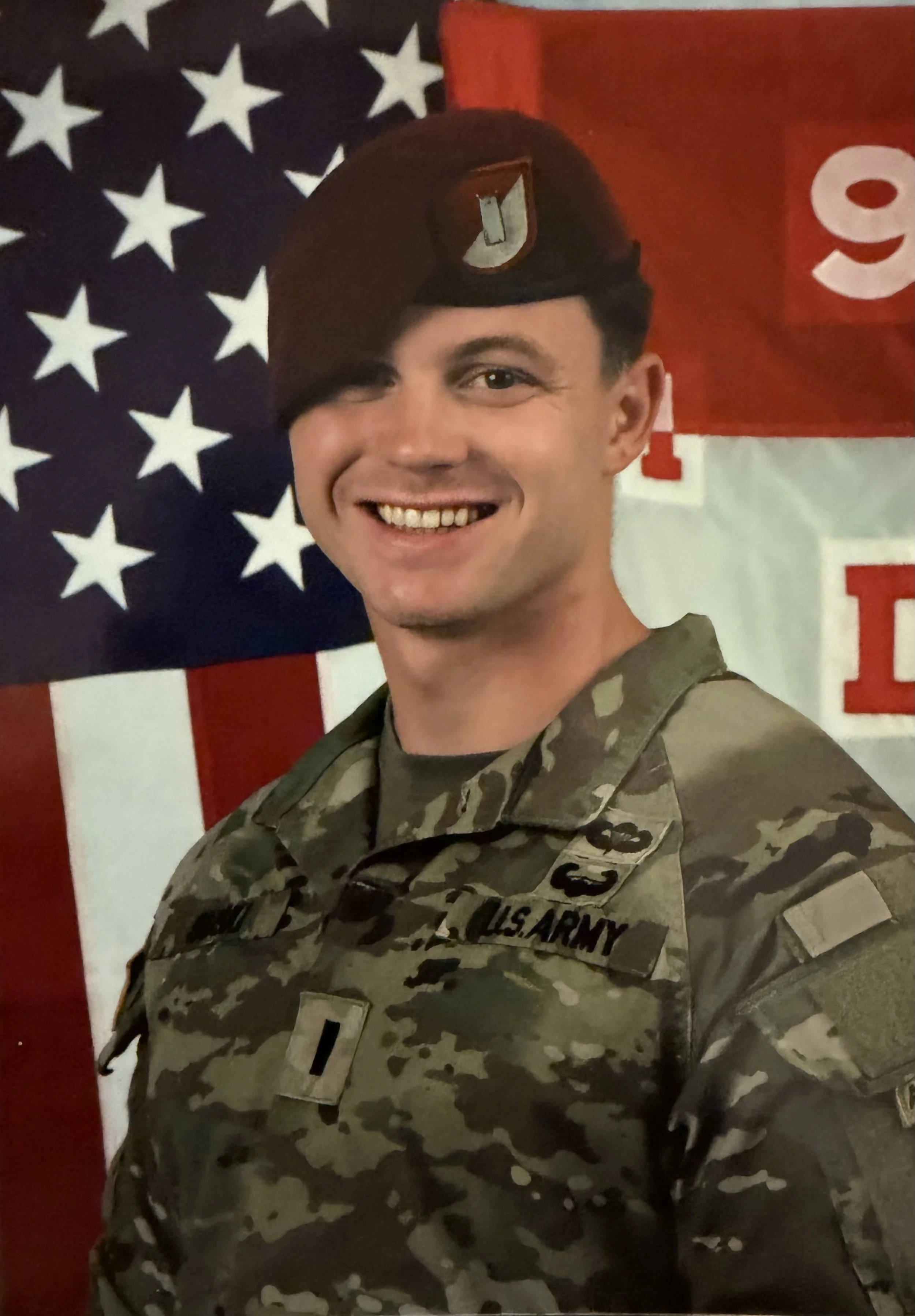 A smiling man in U.S. Army camouflage uniform standing in front of an American flag and a red and white banner.
