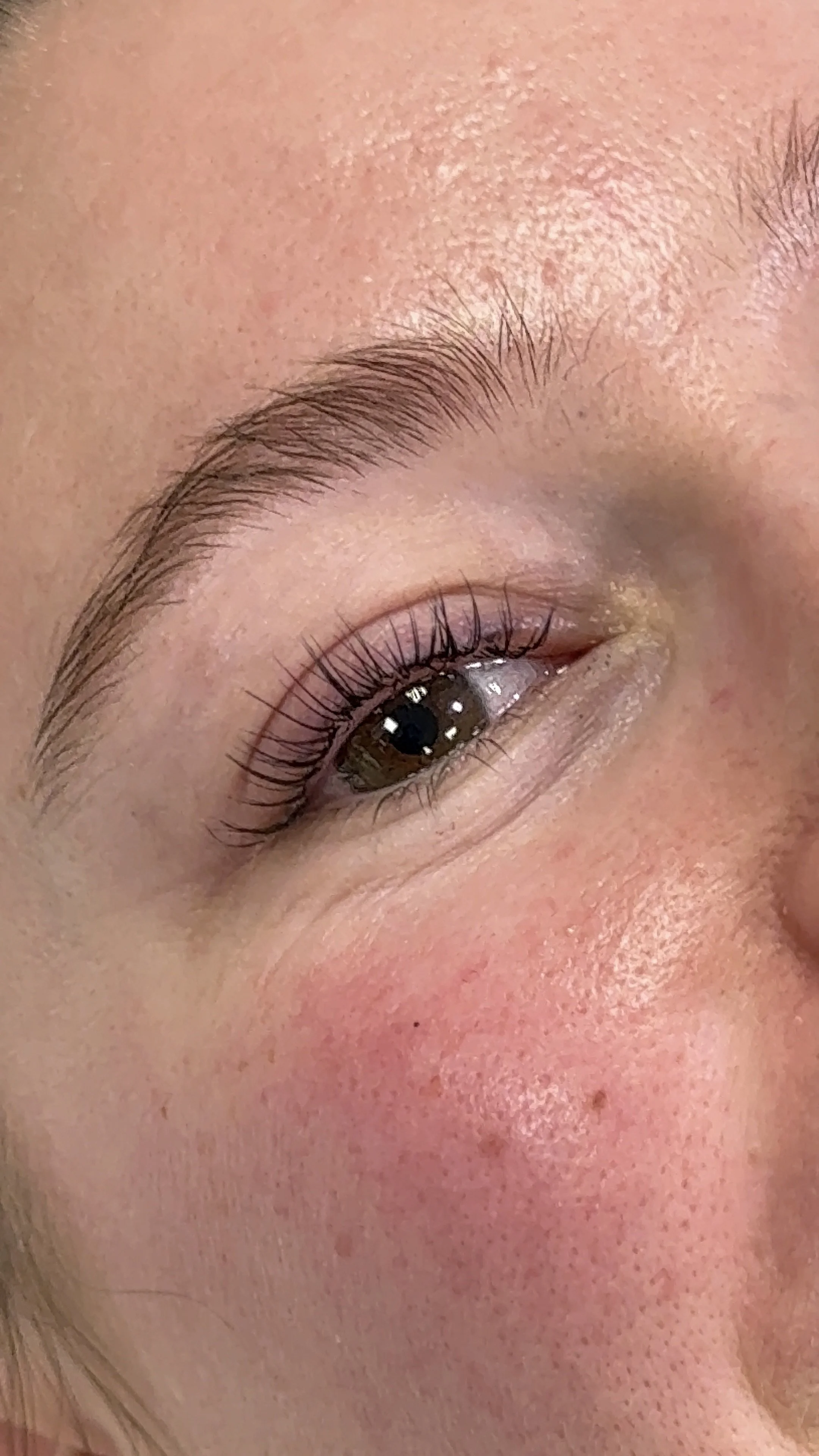 Close-up of a smiling person's face, showing their eye, eyebrow, and part of the cheek, with detailed skin texture and natural makeup.